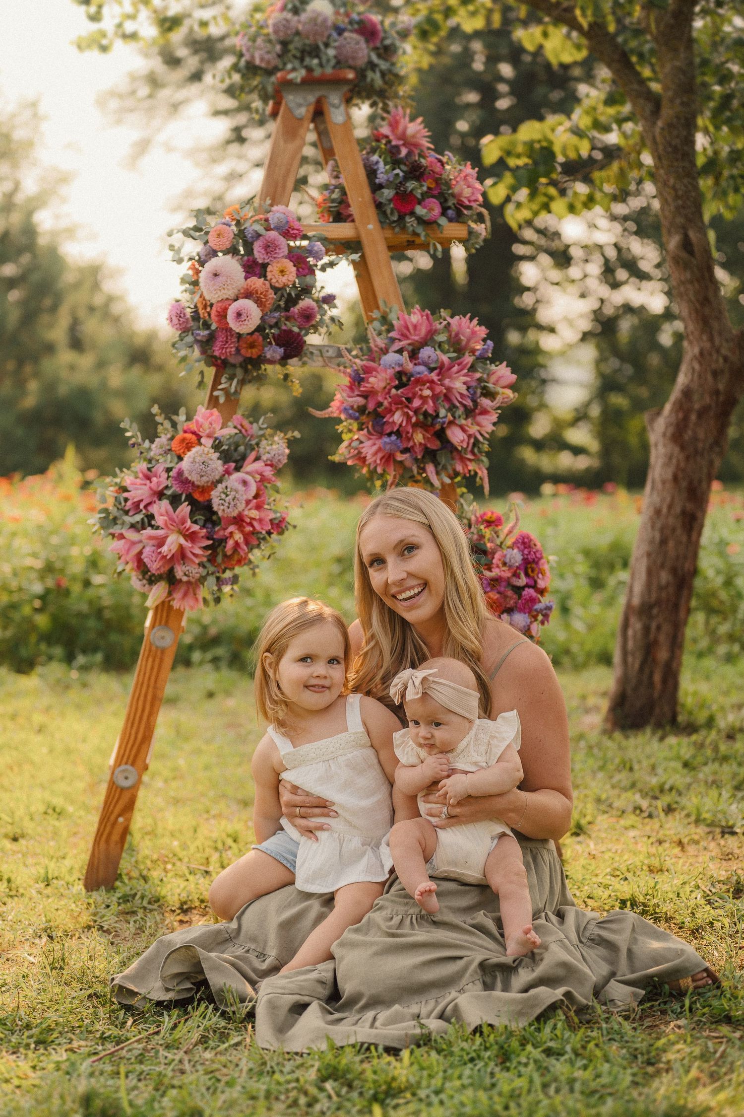 Family sitting on grass near wooden stands with pink and purple floral arrangements at sunset.