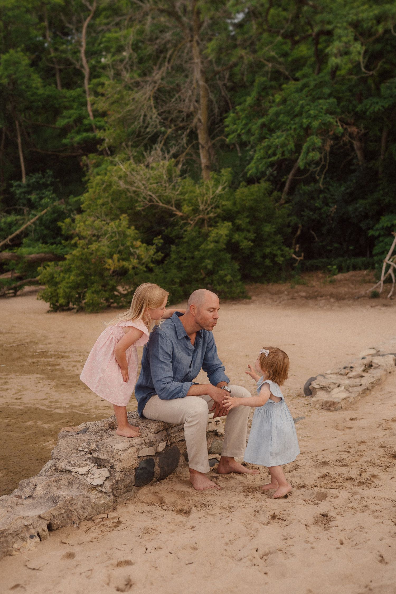 A heartwarming family moment captured on a sandy beach with lush green trees in the background at sunset.