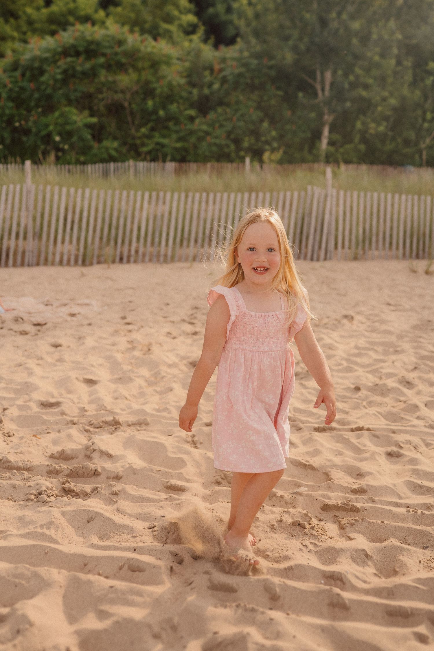 A young child in a pink dress plays happily in the sand at the beach.