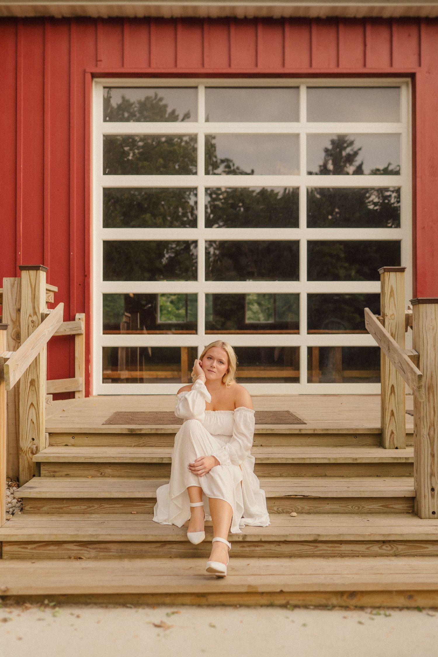 Figure in white clothing sitting on wooden steps of a red building with large windows.