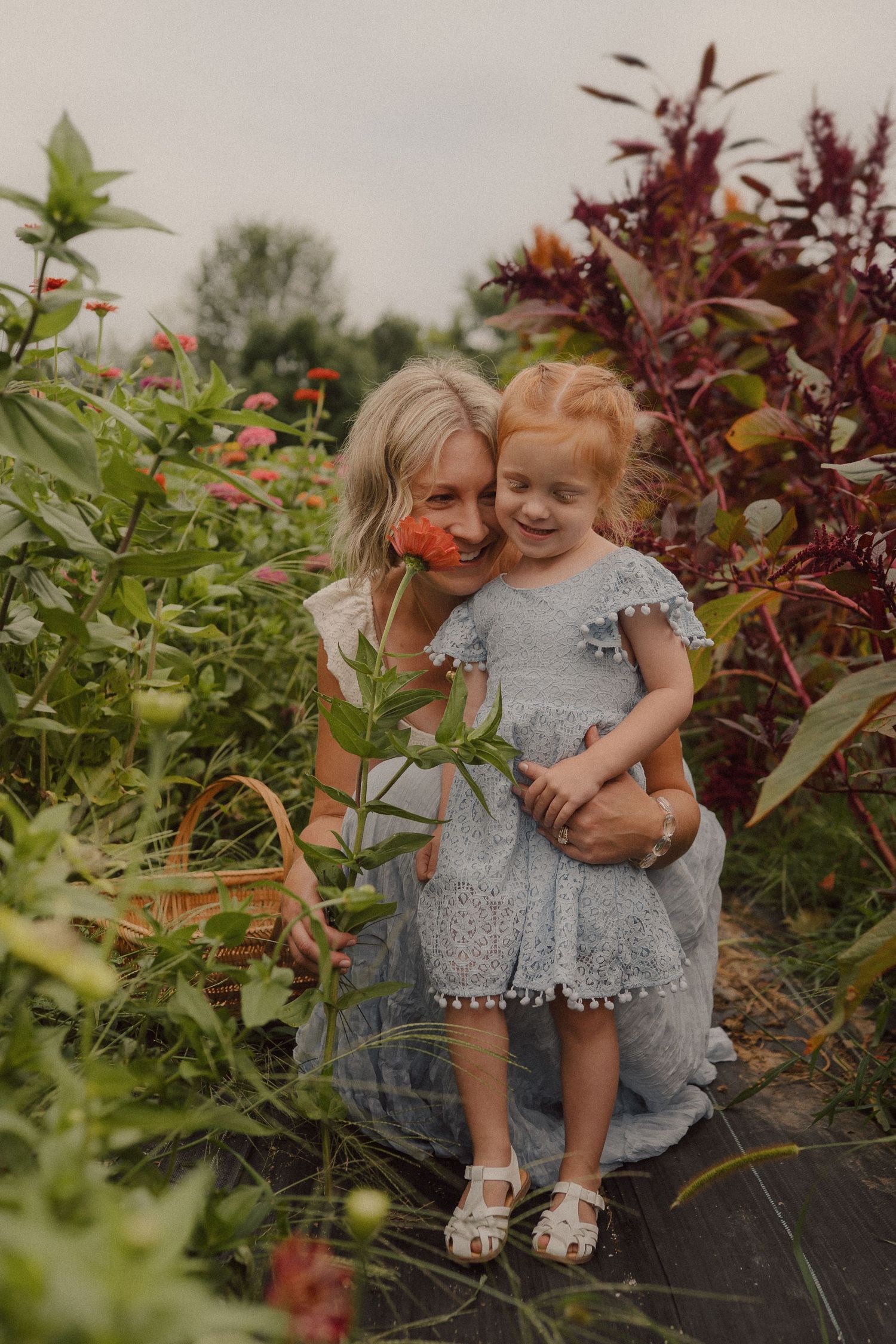 Two children wearing matching gray dresses sit together amid lush garden foliage.