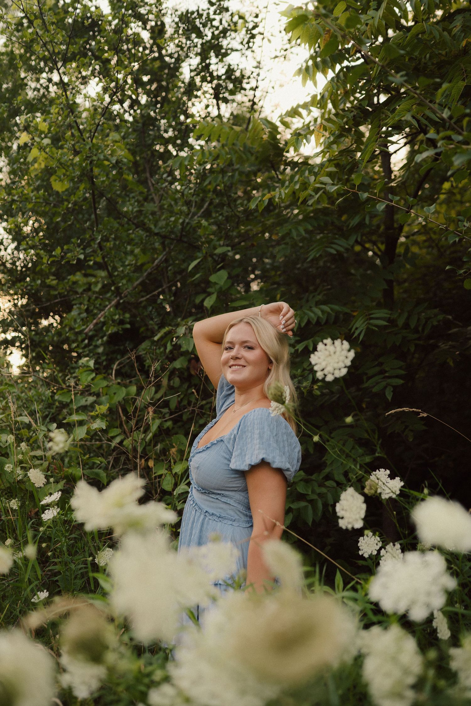 Woman in blue dress poses playfully among white wildflowers and lush green foliage during golden hour.