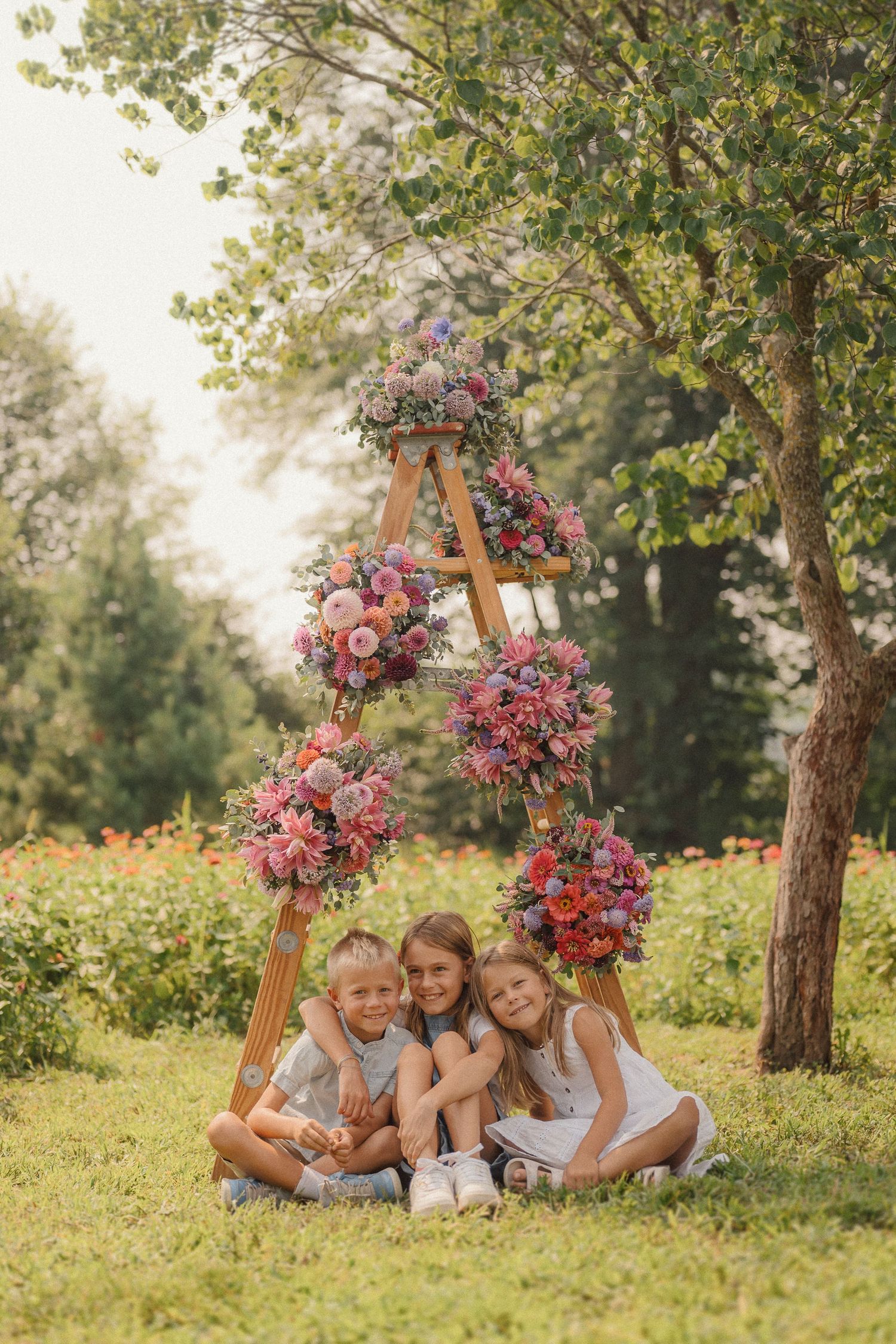 Children sitting together under a wooden ladder decorated with pink and purple floral arrangements in a sunny garden.