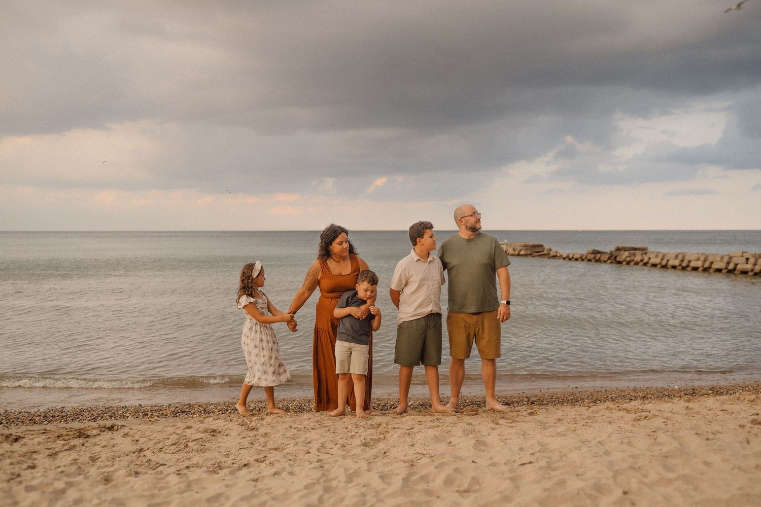 A family stands together on a sandy beach at sunset with stormy clouds overhead and a jetty in the background.