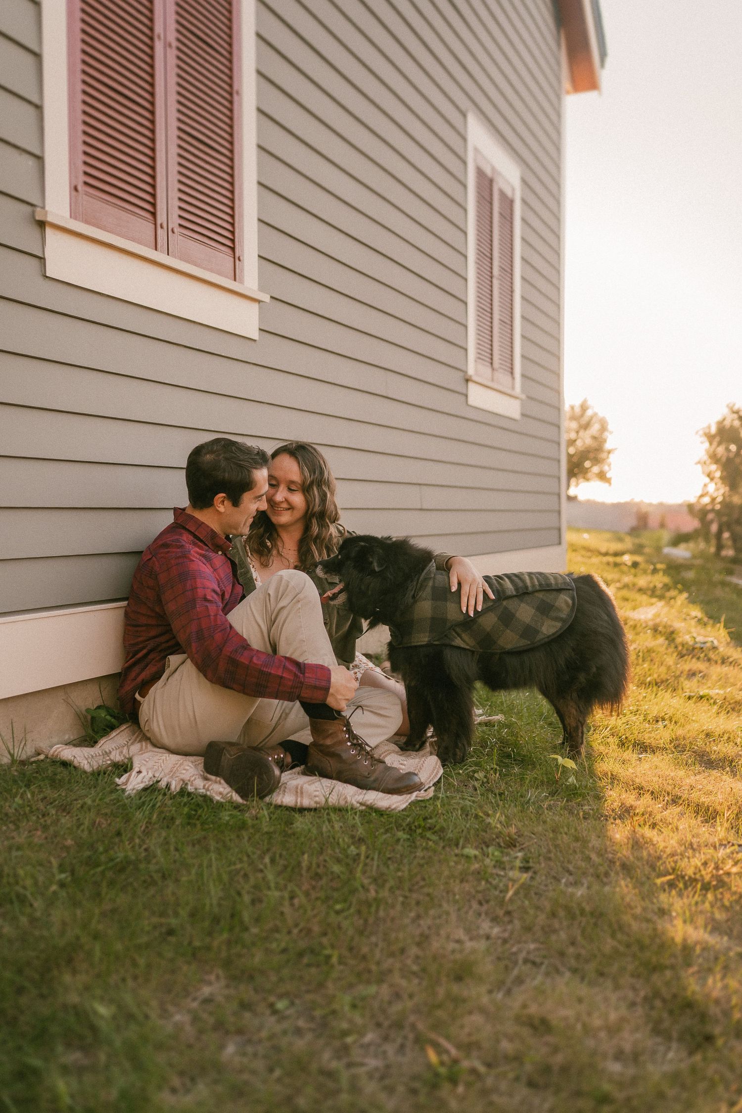 A couple sits with their black dog beside a grey house in warm evening light.