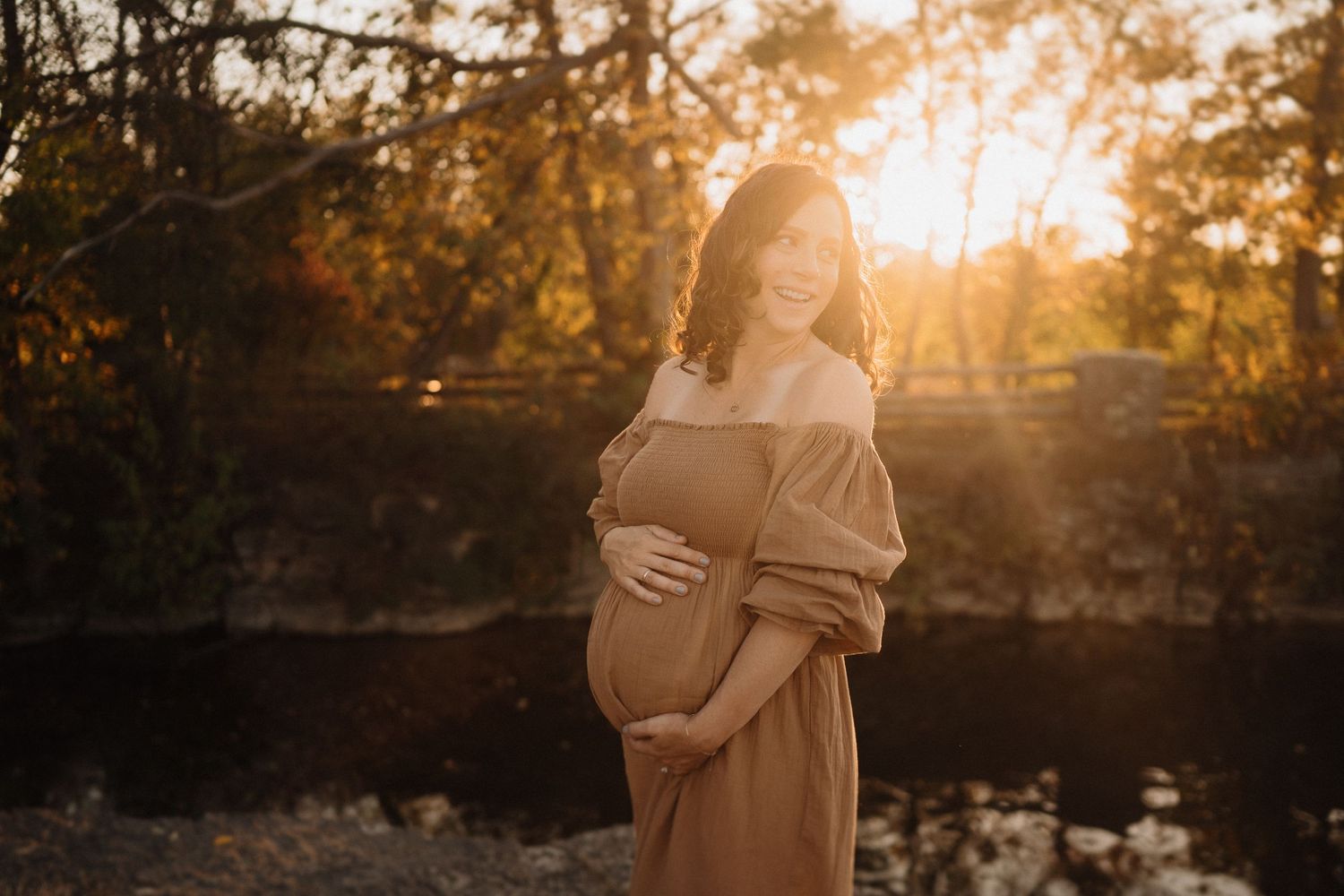Warm sunset maternity portrait shows a woman in a flowy brown dress cradling her baby bump.