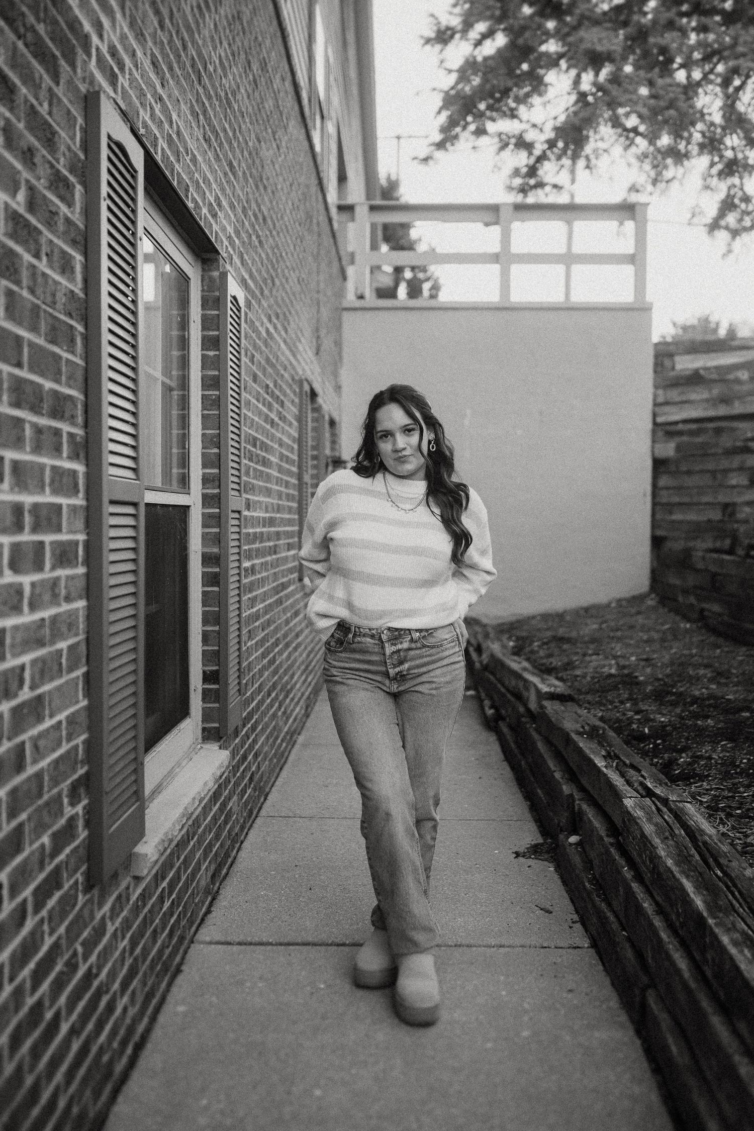 Person in striped sweater and jeans stands on brick walkway beside building in black and white photo.