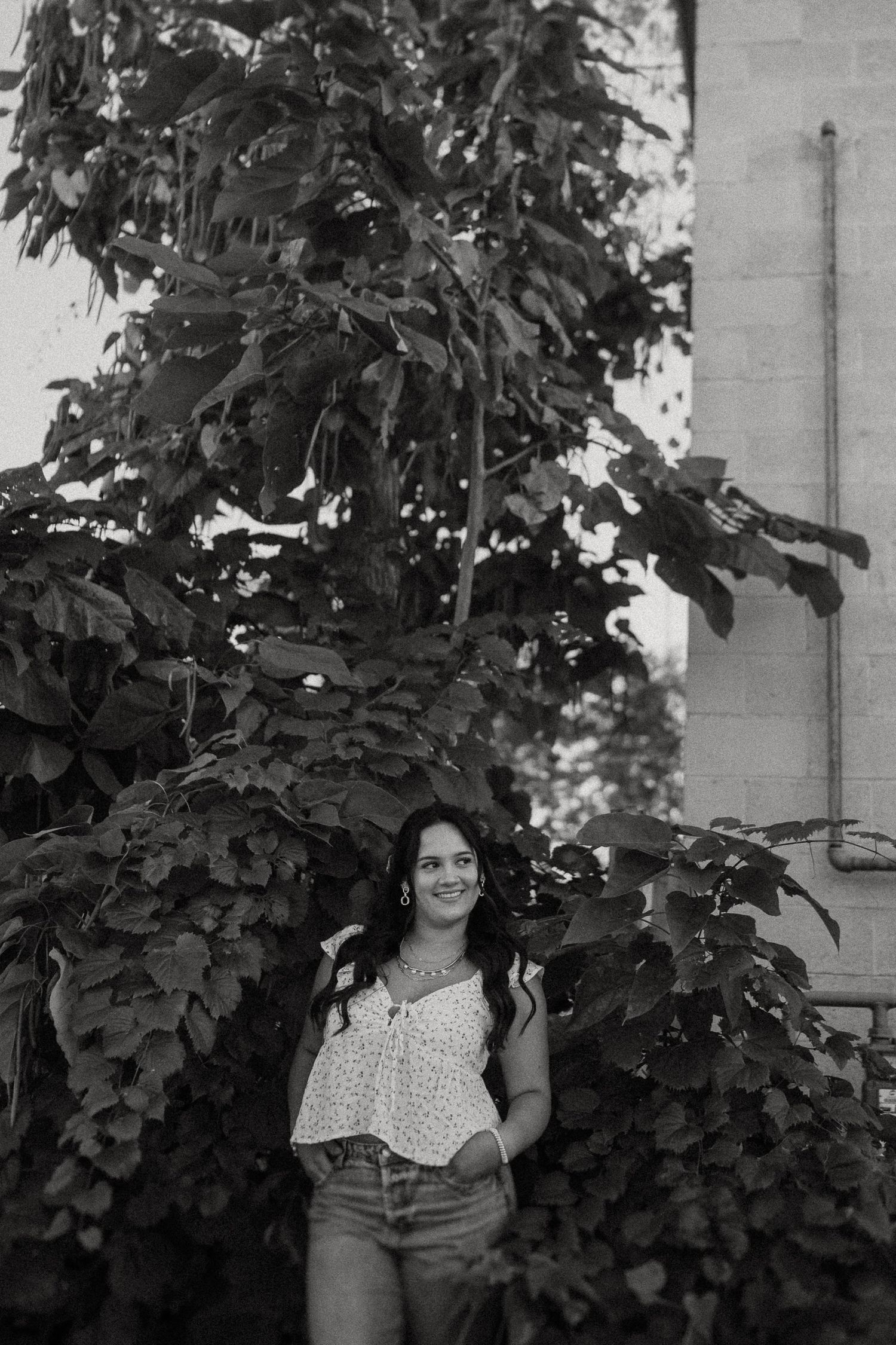 A black and white portrait of someone standing next to leafy foliage while wearing polka dot top and jeans.