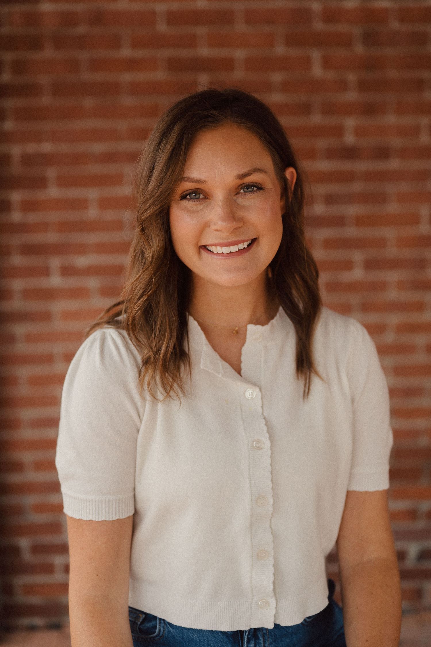 Headshot portrait of someone wearing a white short-sleeved top against a brick wall background.