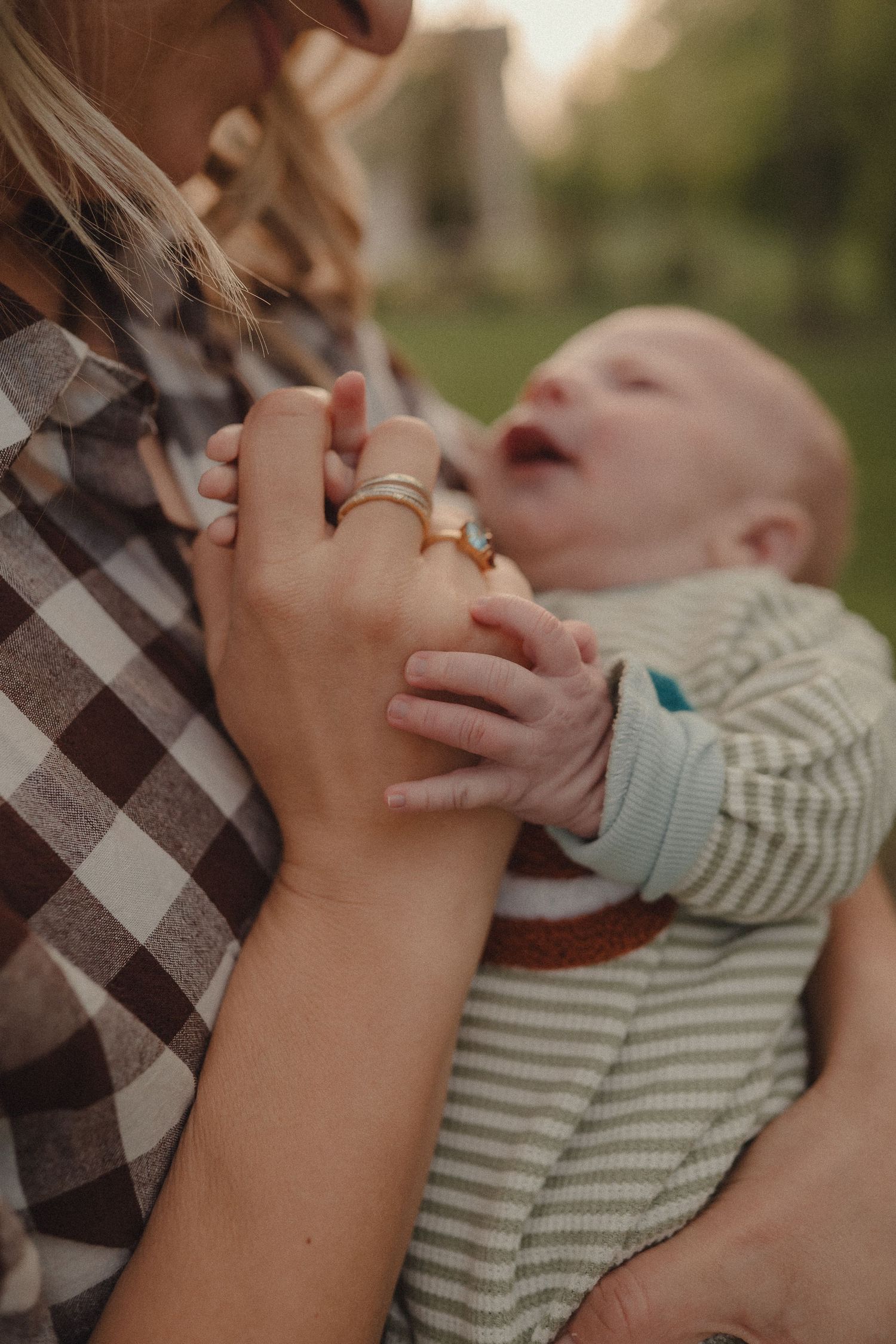 A mother holds her newborn baby who grips her finger outdoors in soft golden light.