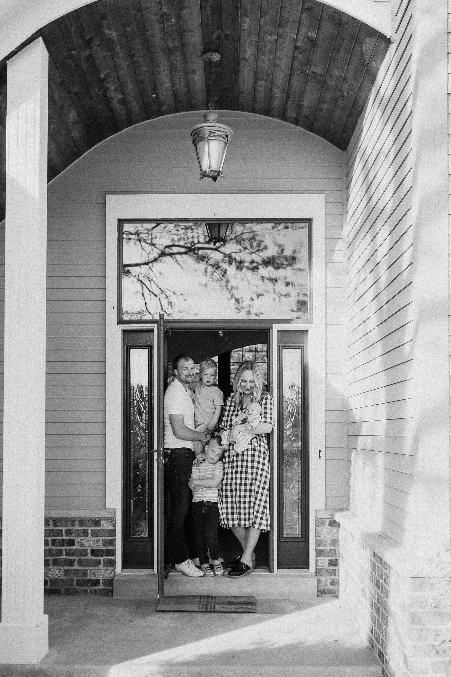 Black and white photo of a young family with two children standing in their home's front doorway under an arched porch.