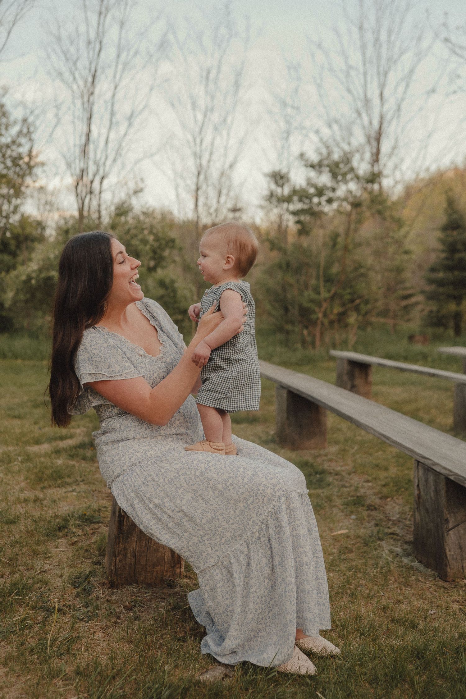 Smiling mother in gray floral dress sitting on log bench holding standing baby in green outfit in peaceful park setting.