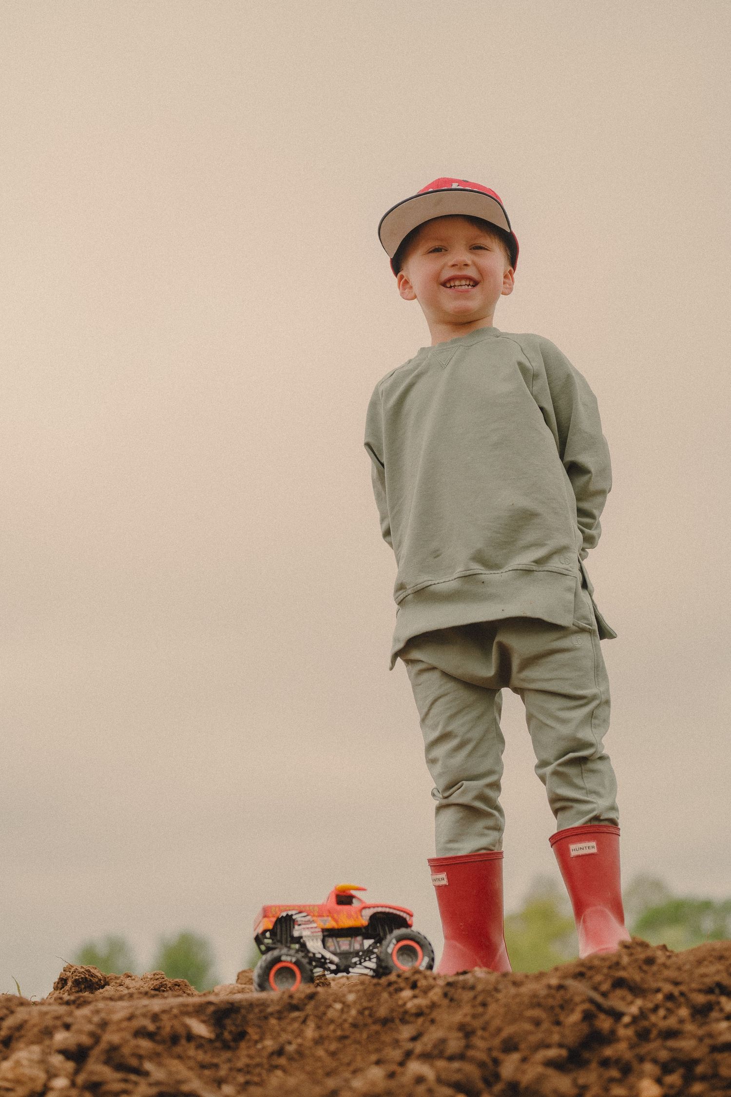 Smiling boy in green outfit and red boots stands on dirt mound with toy monster truck.