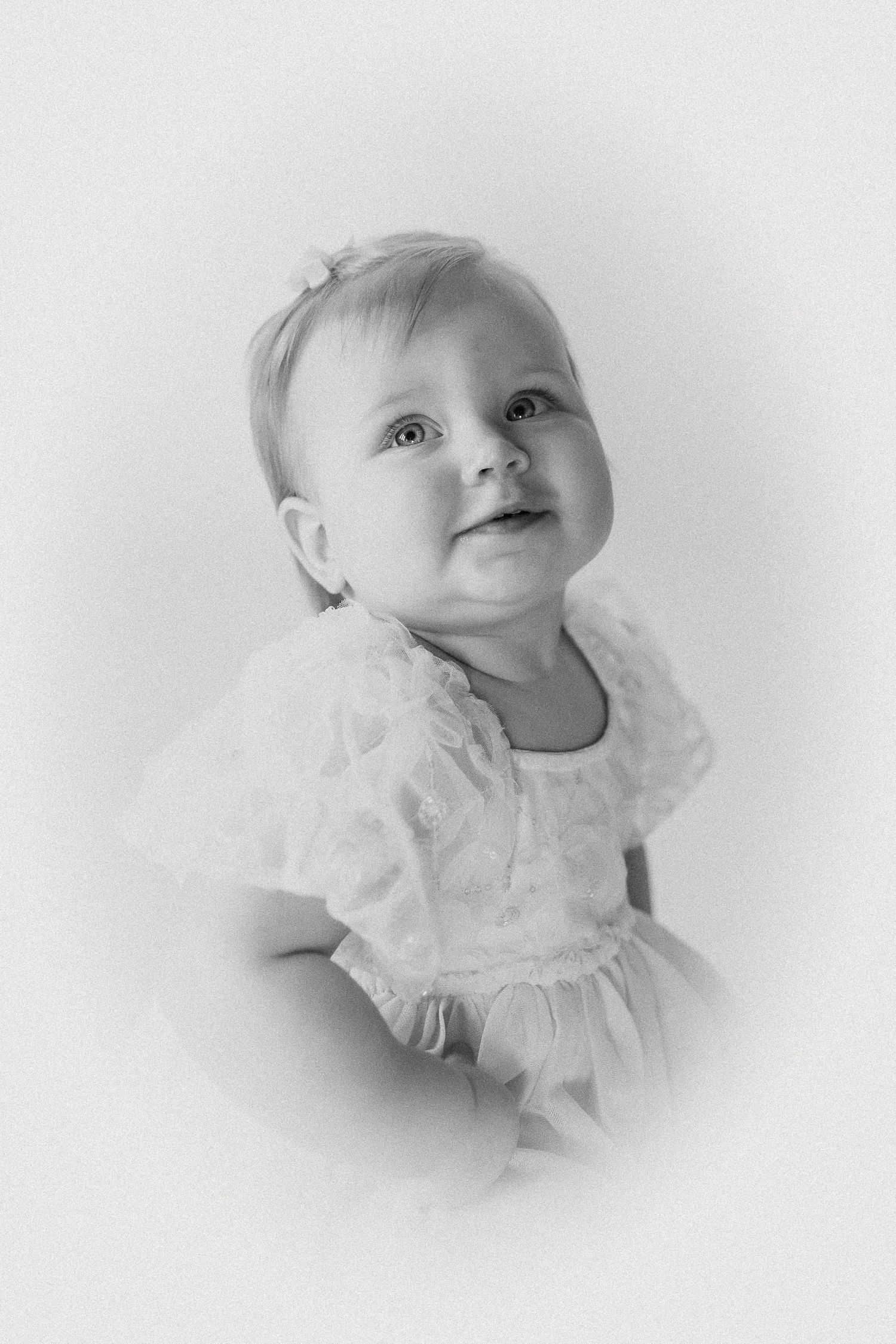 Soft black and white portrait of an adorable baby girl in a lace dress gazing upward with a gentle smile.