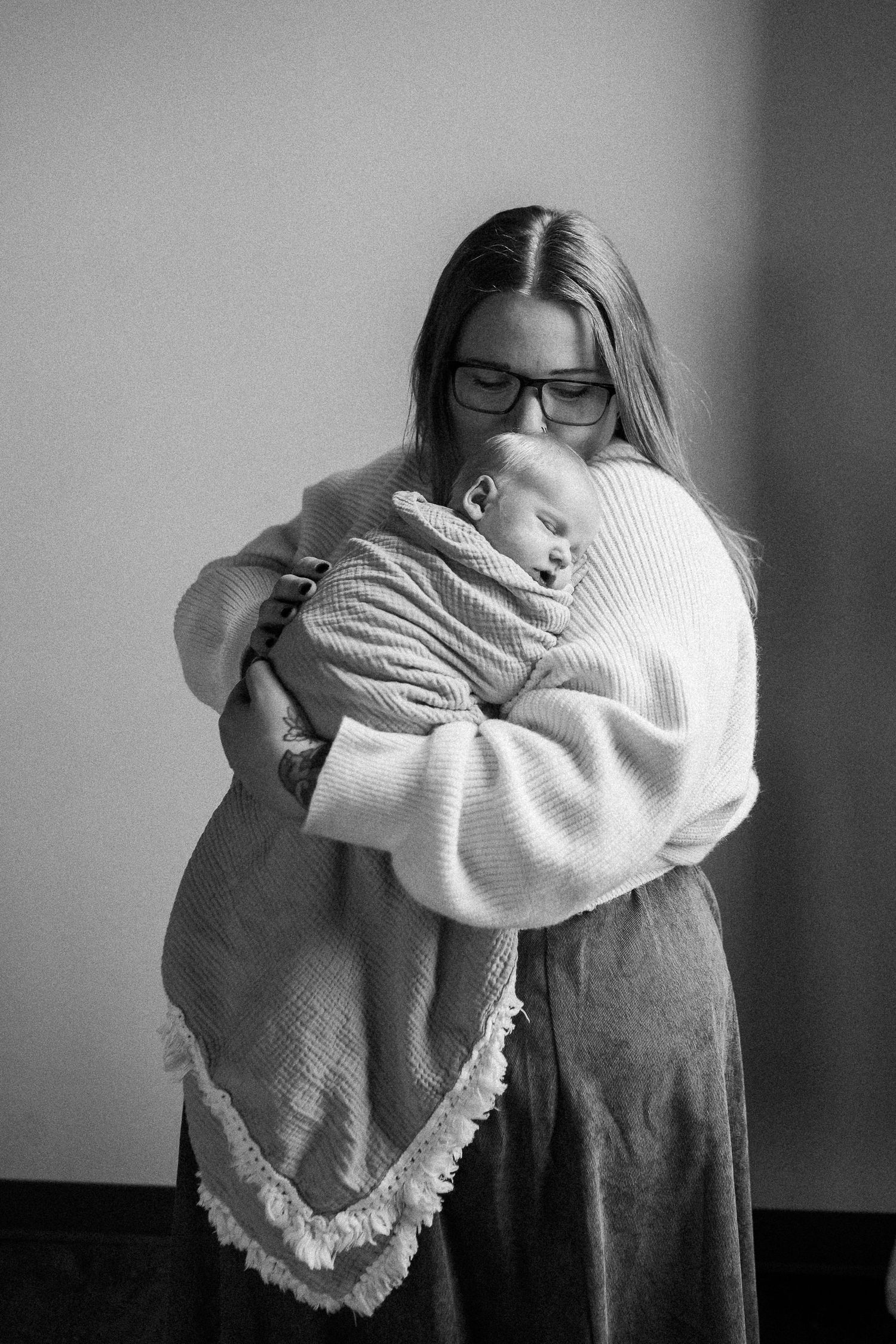 Mother in glasses tenderly holding sleeping newborn baby wrapped in white blanket, black and white photo.