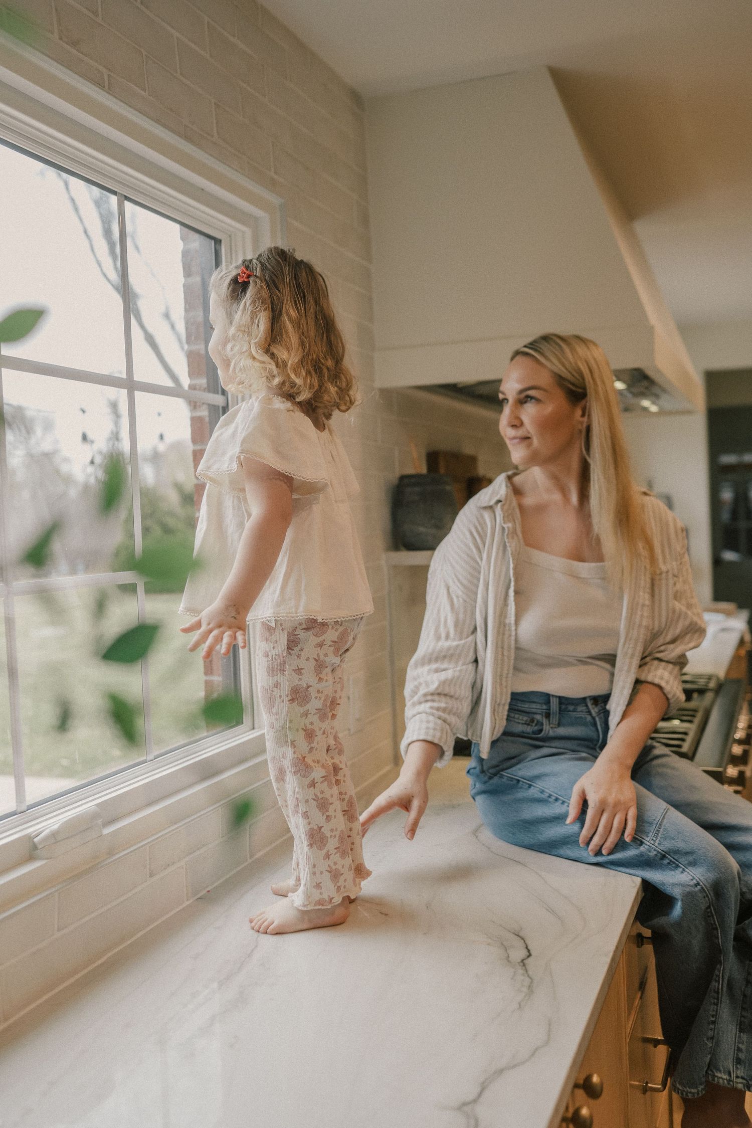 A blonde mother sits on a marble kitchen counter watching her toddler daughter stand and look out the window.