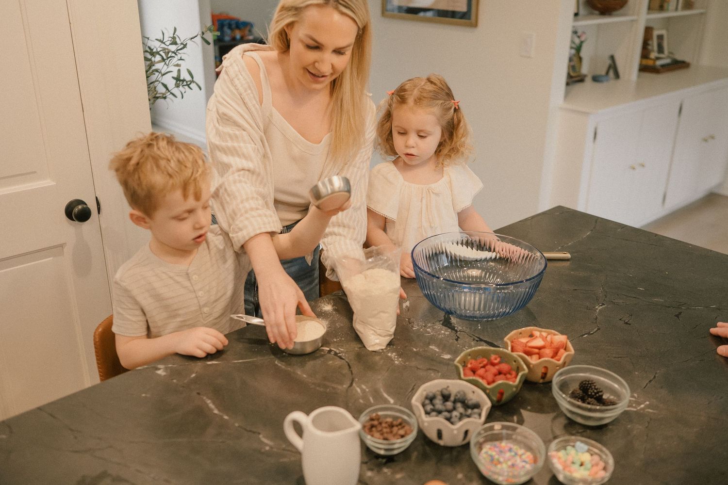 A mother and two young children baking together at a kitchen counter with colorful sprinkles and mixing bowls.