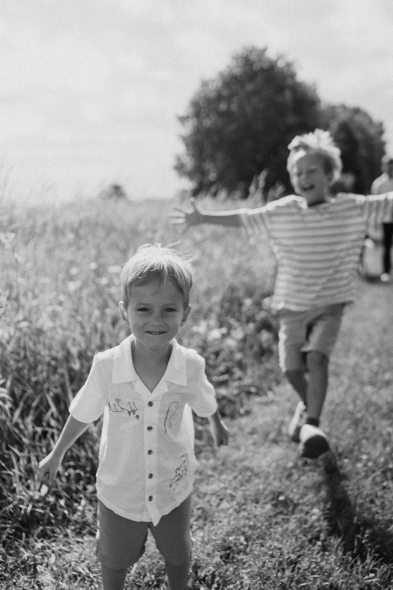 Two joyful boys running through a sunny green field, laughing and playing outdoors in a black and white photo.