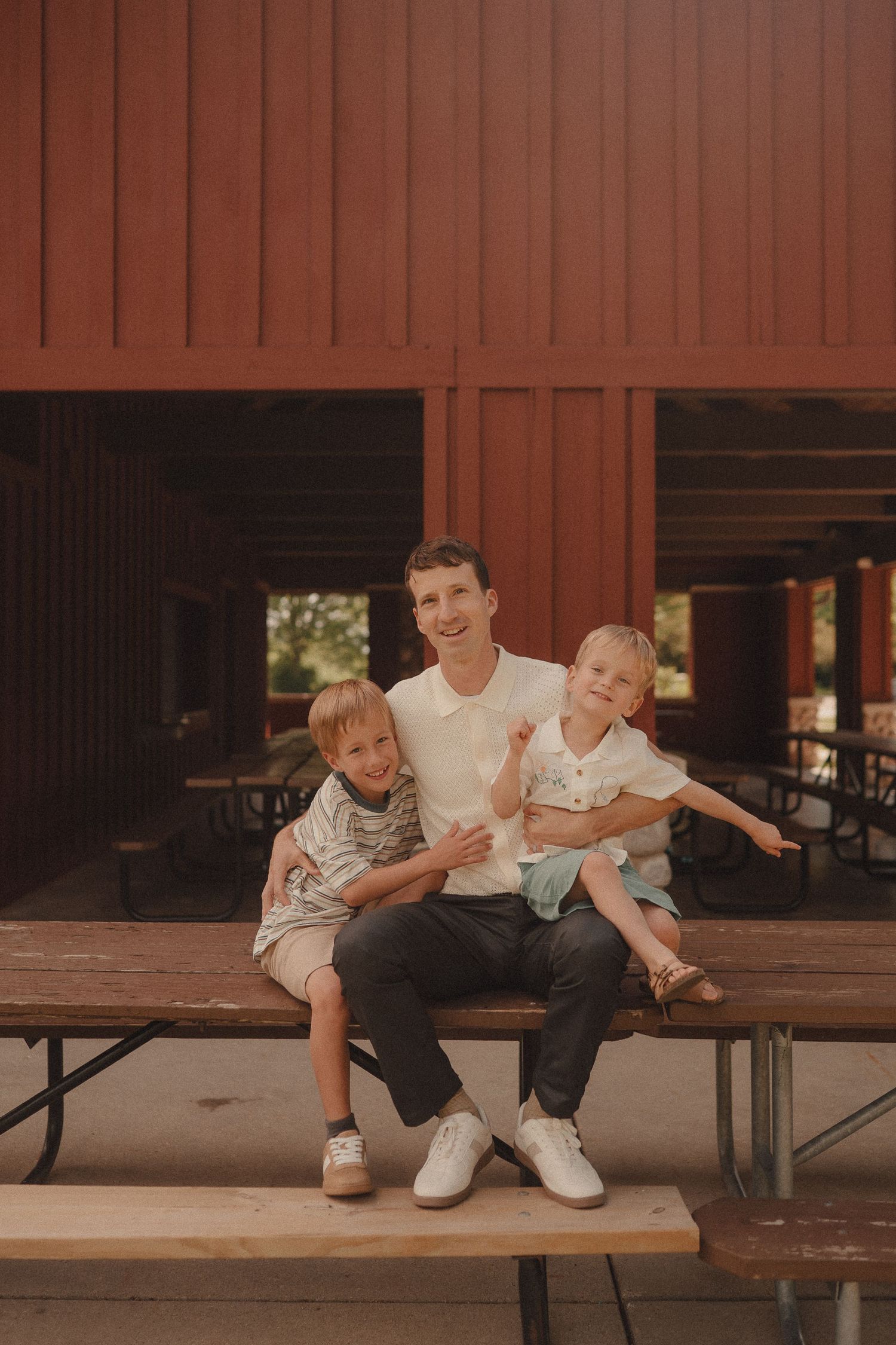 A smiling father sits on a wooden bench with his two young sons in front of a red building.