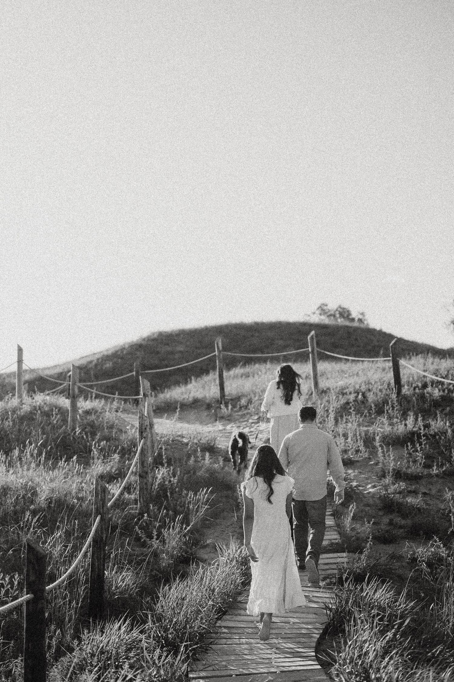 Family walking up a wooden boardwalk path through grassy hills in black and white photography.