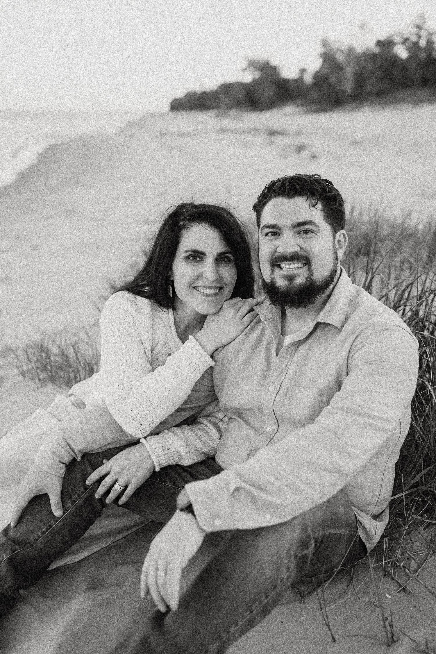 Black and white beach portrait of smiling couple sitting together on sandy shore.
