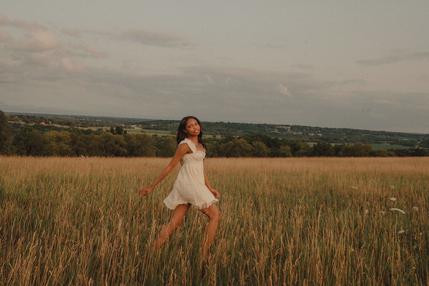 Young woman in a white dress walking through a golden field at dusk with scenic countryside views.