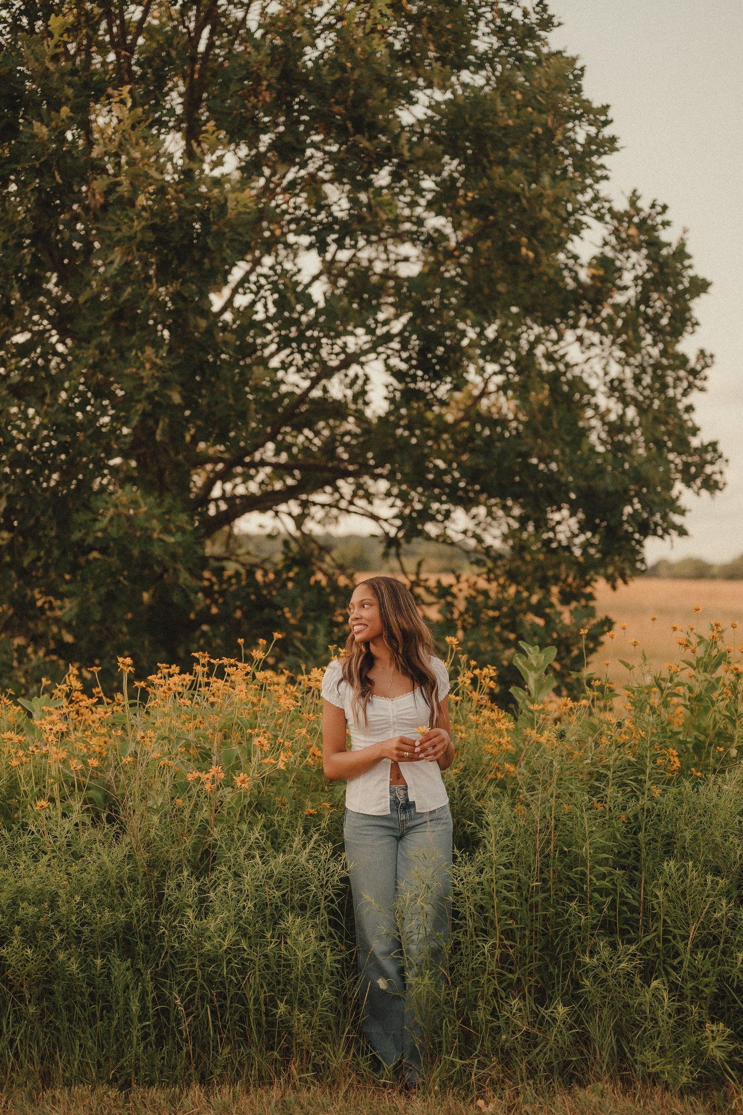 Smiling young woman standing among yellow wildflowers near a large oak tree at sunset.