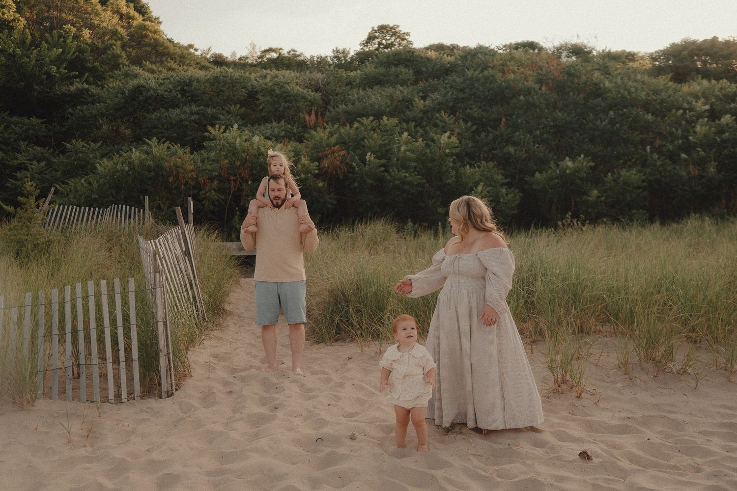 Family of four enjoying a beach walk on sandy dunes with lush greenery, mom in flowing dress, dad with child on shoulders.