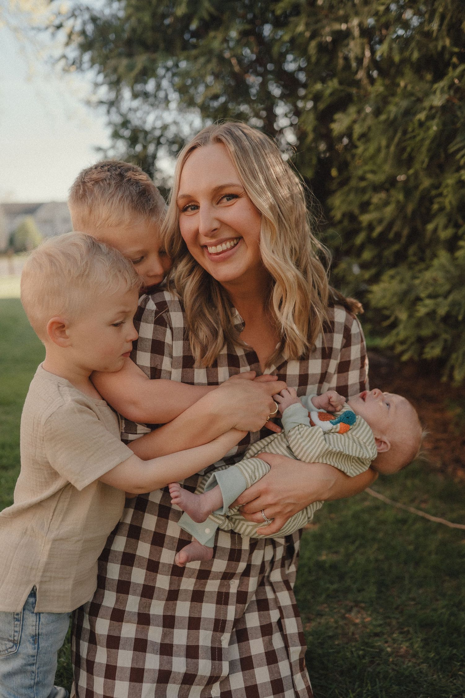 A smiling blonde mother holds a newborn while two young boys hug her outdoors near evergreen trees.