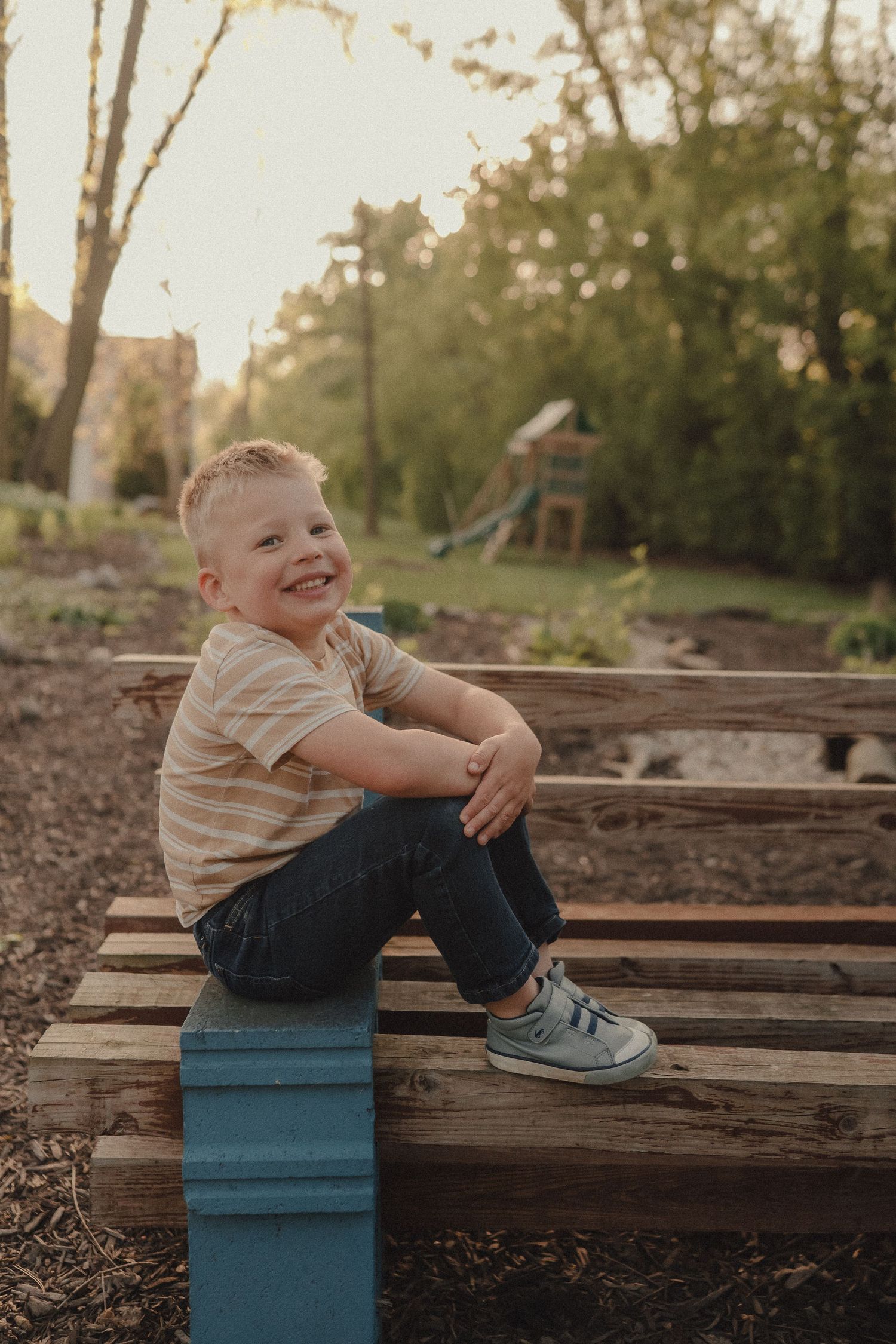A smiling young boy sits on wooden pallets in a backyard garden at golden hour.