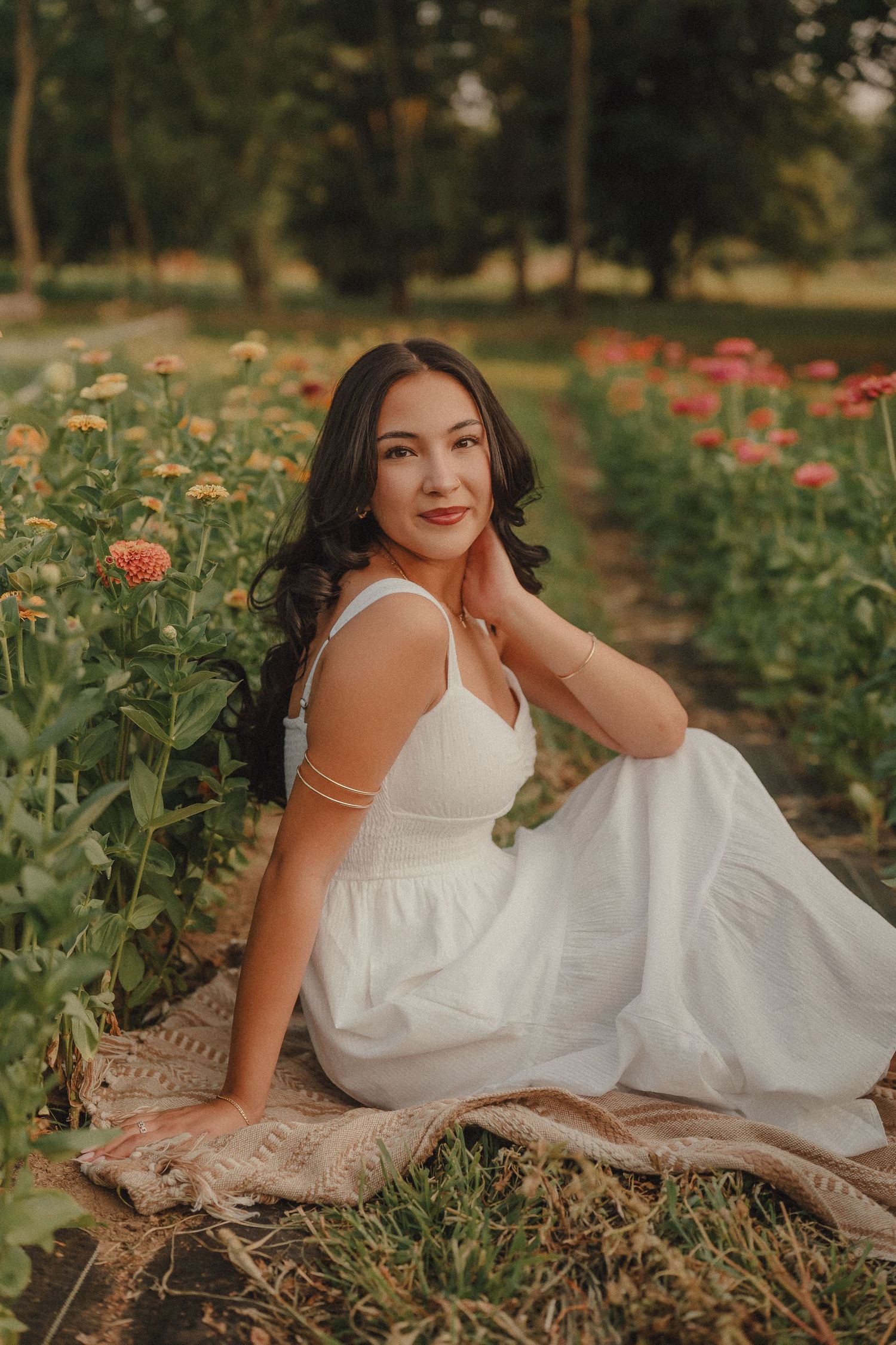 Woman in white dress sitting gracefully among colorful zinnia flowers at sunset.
