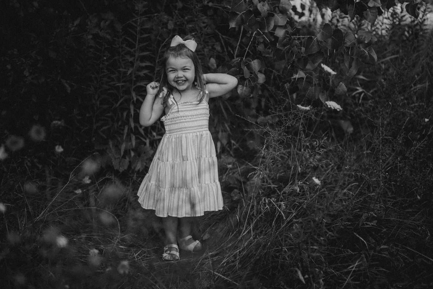 A black and white portrait of a child in a dress playing outdoors among wild plants.