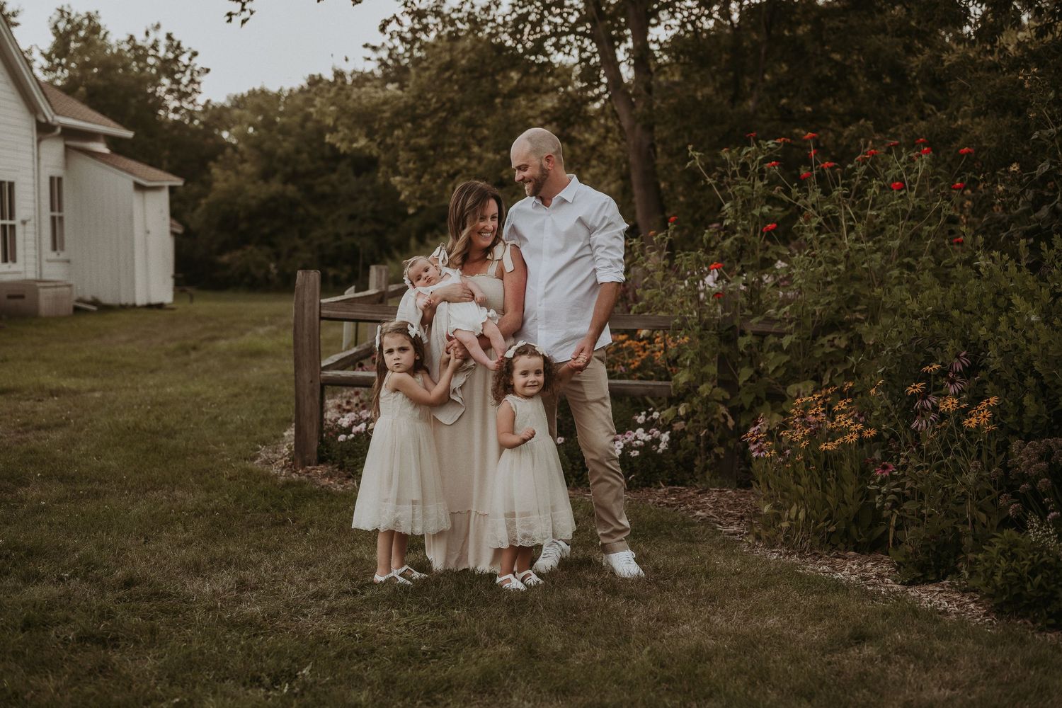 Family in coordinated cream outfits poses near blooming roses and a white farmhouse.