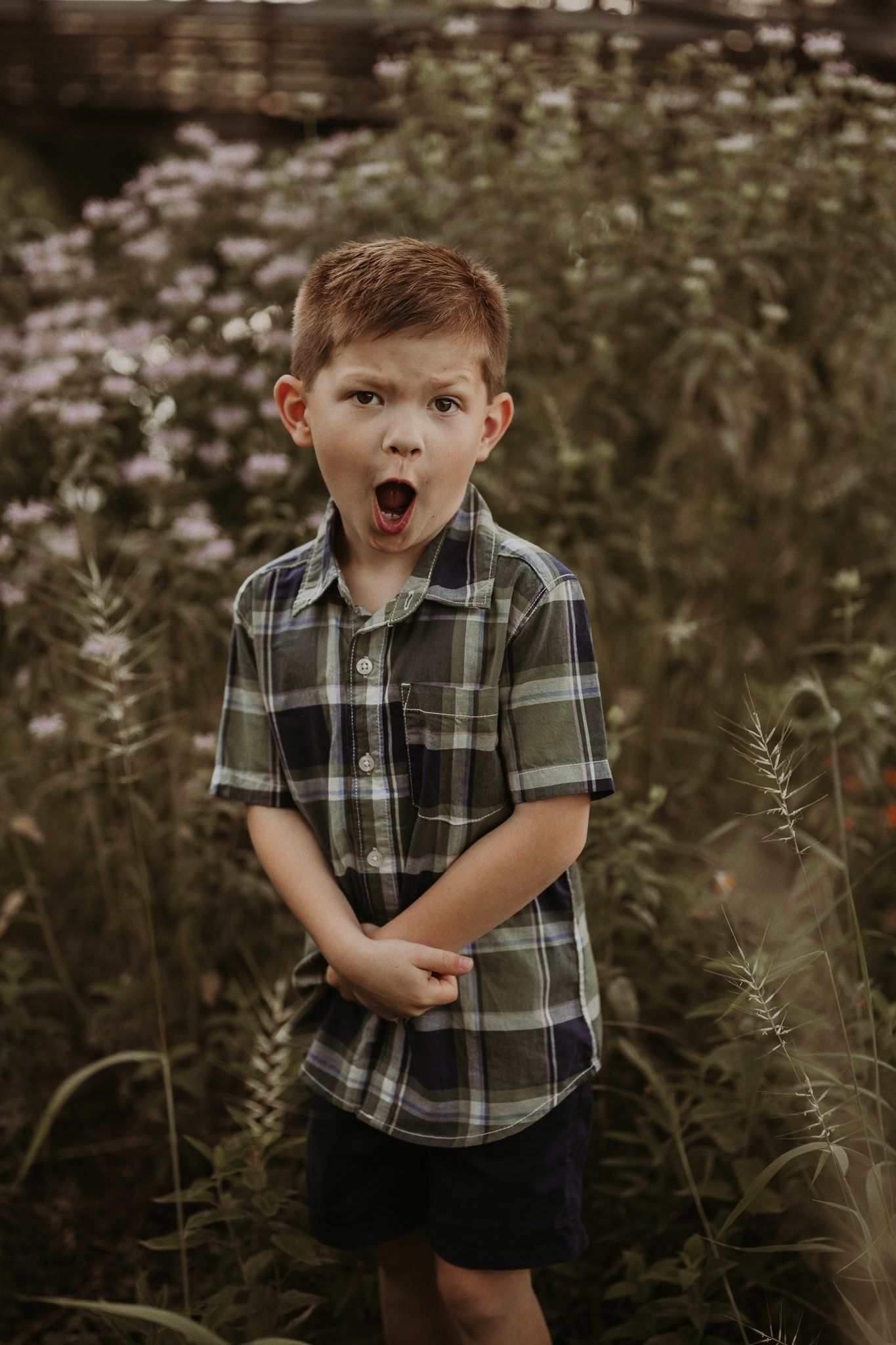 Young child in plaid shirt shows surprised expression while standing in wildflower field at sunset.