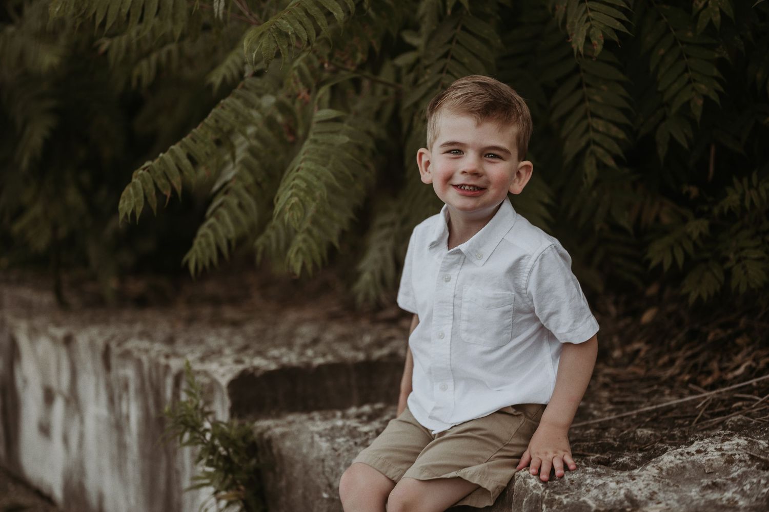 A happy child wearing khaki shorts sits casually on stone wall outdoors.