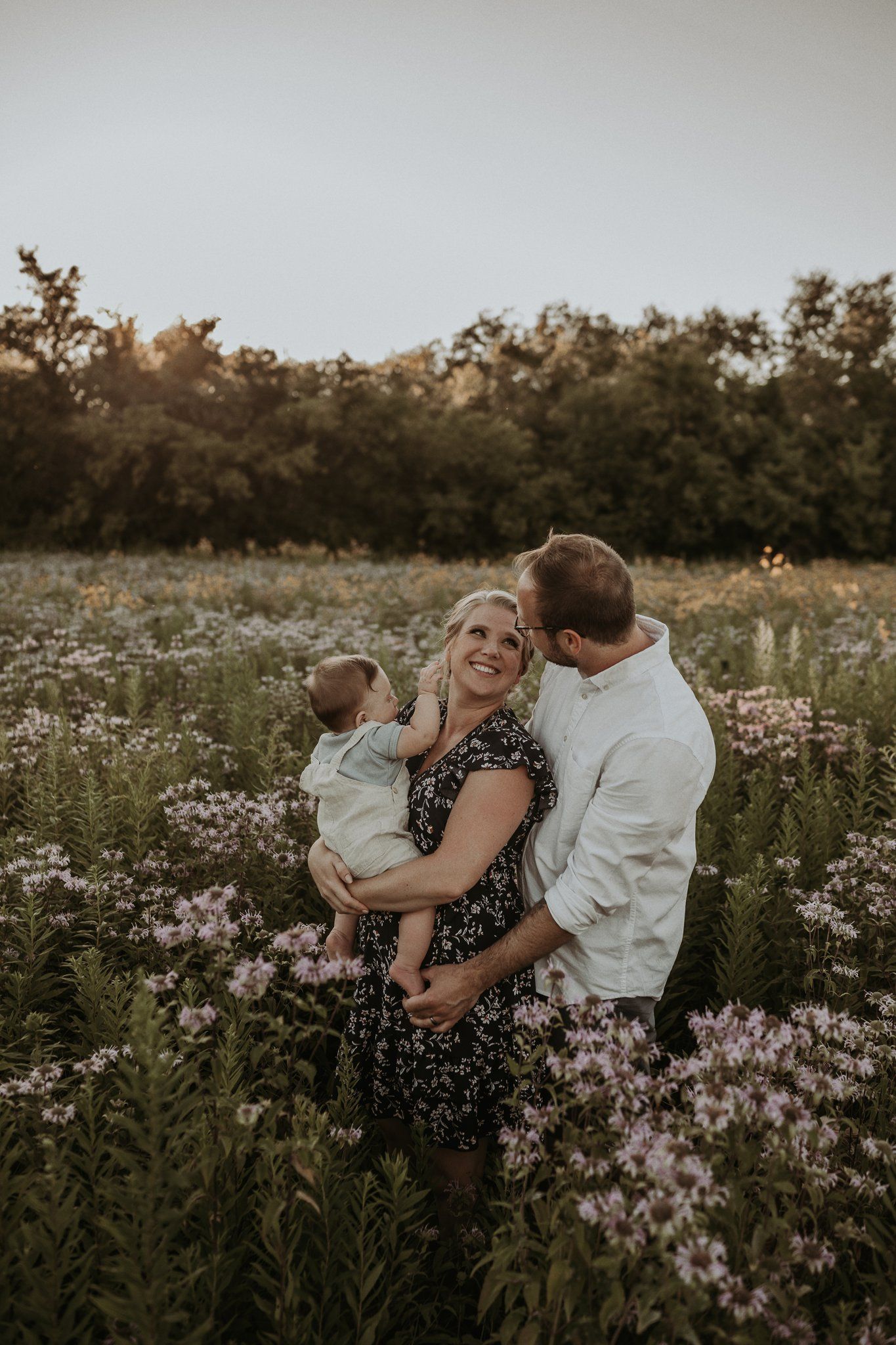 A family embraces warmly in a blooming wildflower field during golden hour with trees in background.