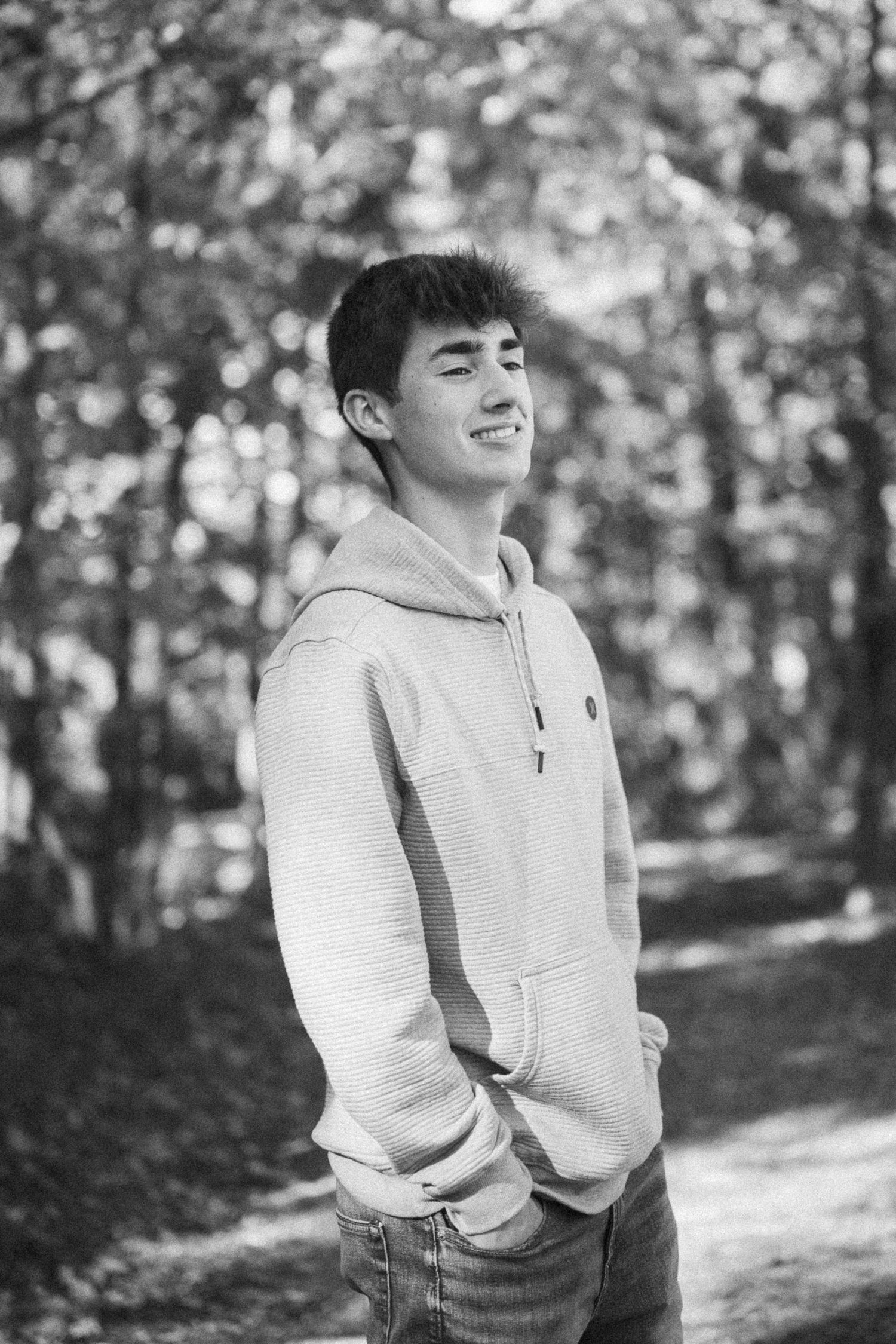 Black and white portrait of someone in a light-colored sweatshirt smiling against a blurred forest backdrop.