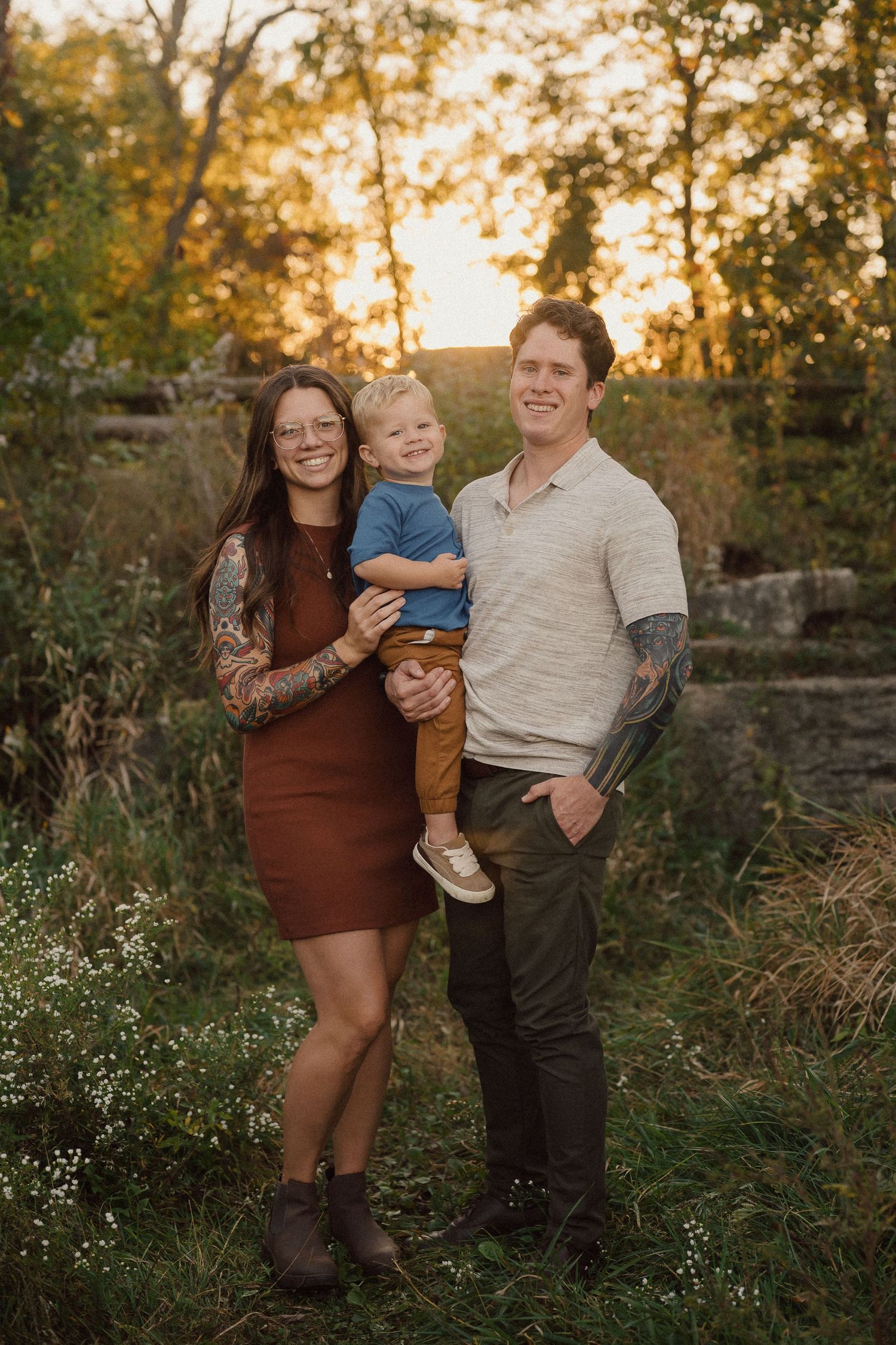 Family of three posing together in autumn setting with golden sunlight filtering through trees.