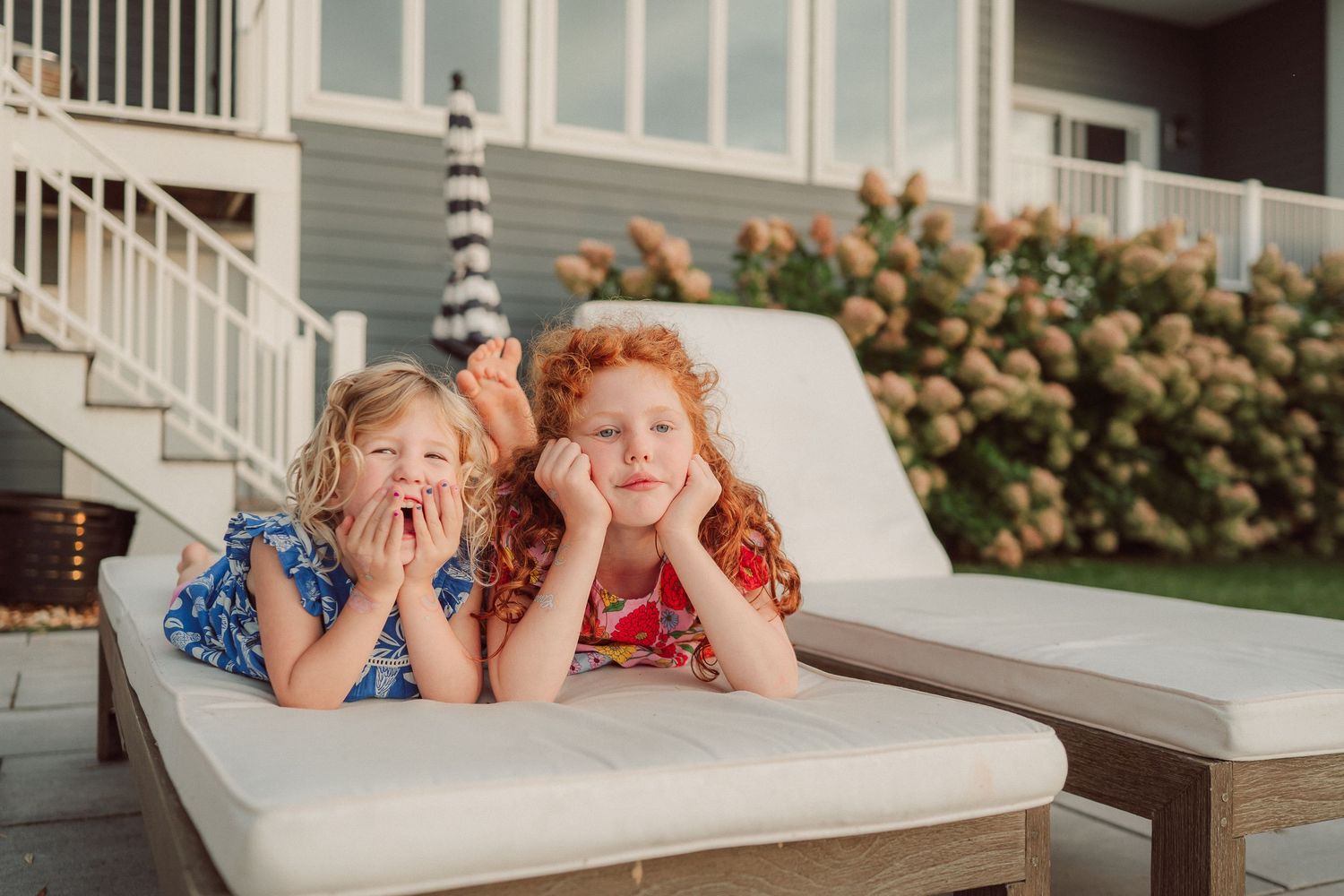 Two children pose playfully on an outdoor chaise lounge with hydrangea bushes in the background.