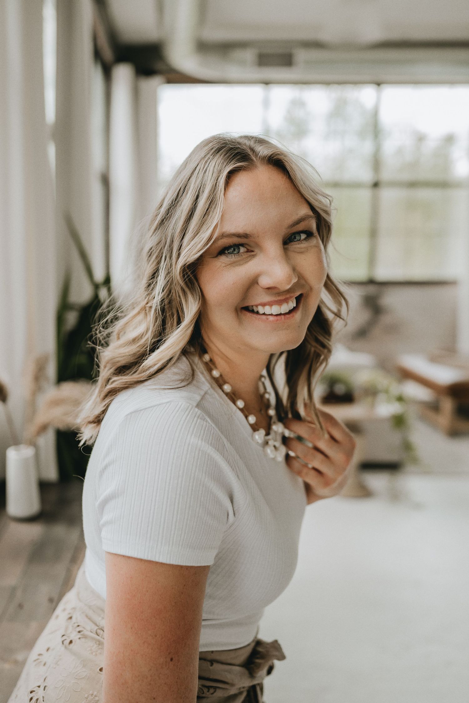 Woman in white t-shirt and pearl necklace smiles brightly in sunlit room.