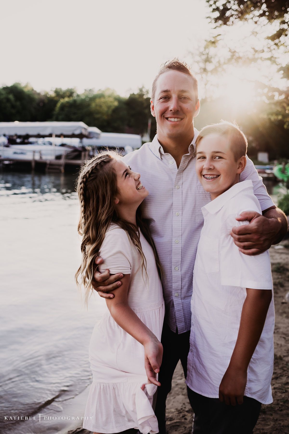 Three people embrace and laugh together by a marina during golden hour.