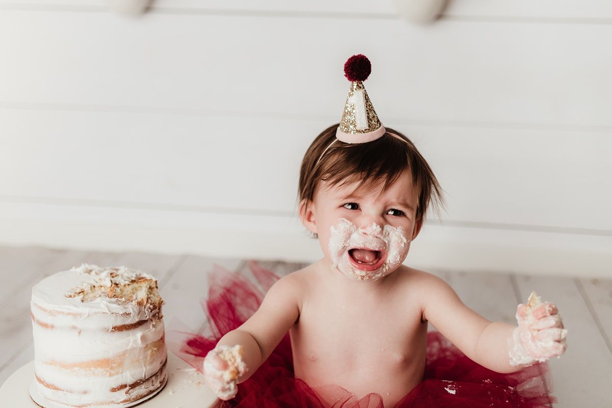 Laughing baby wearing party hat celebrates with smashed cake and red tulle.