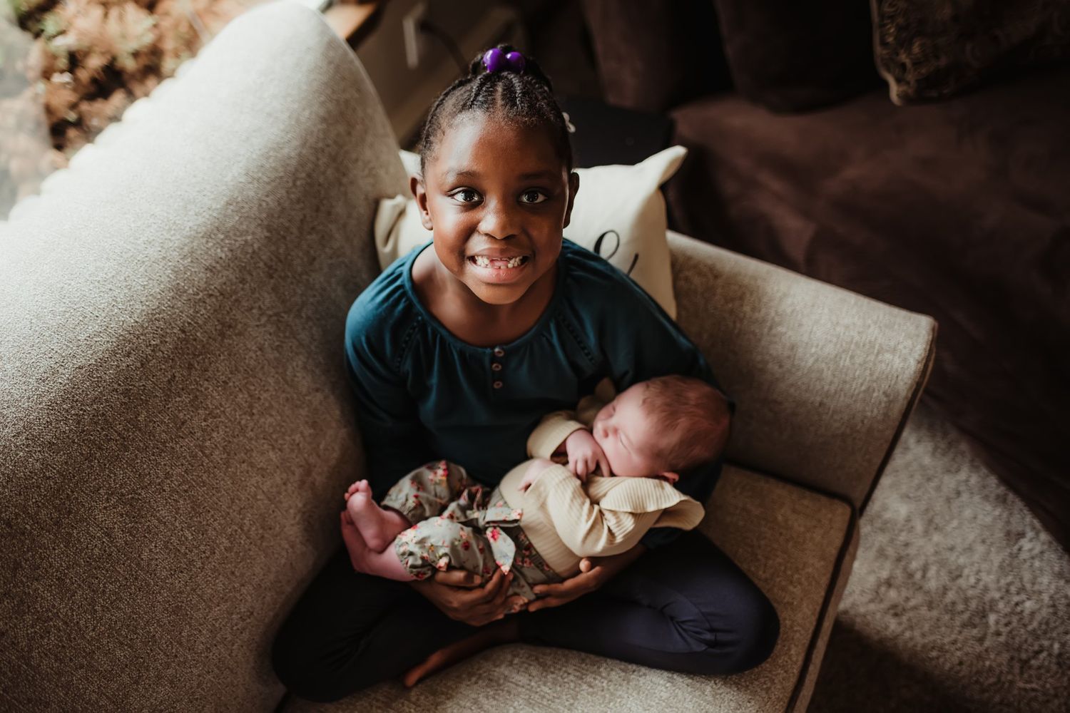 Young child on couch lovingly cradling and gazing at a sleeping newborn.