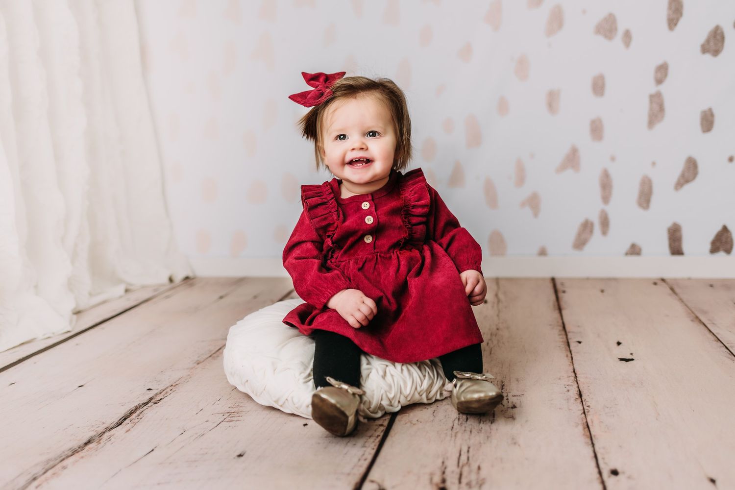 Baby in red ruffled dress sitting on wooden floor with spotted backdrop.