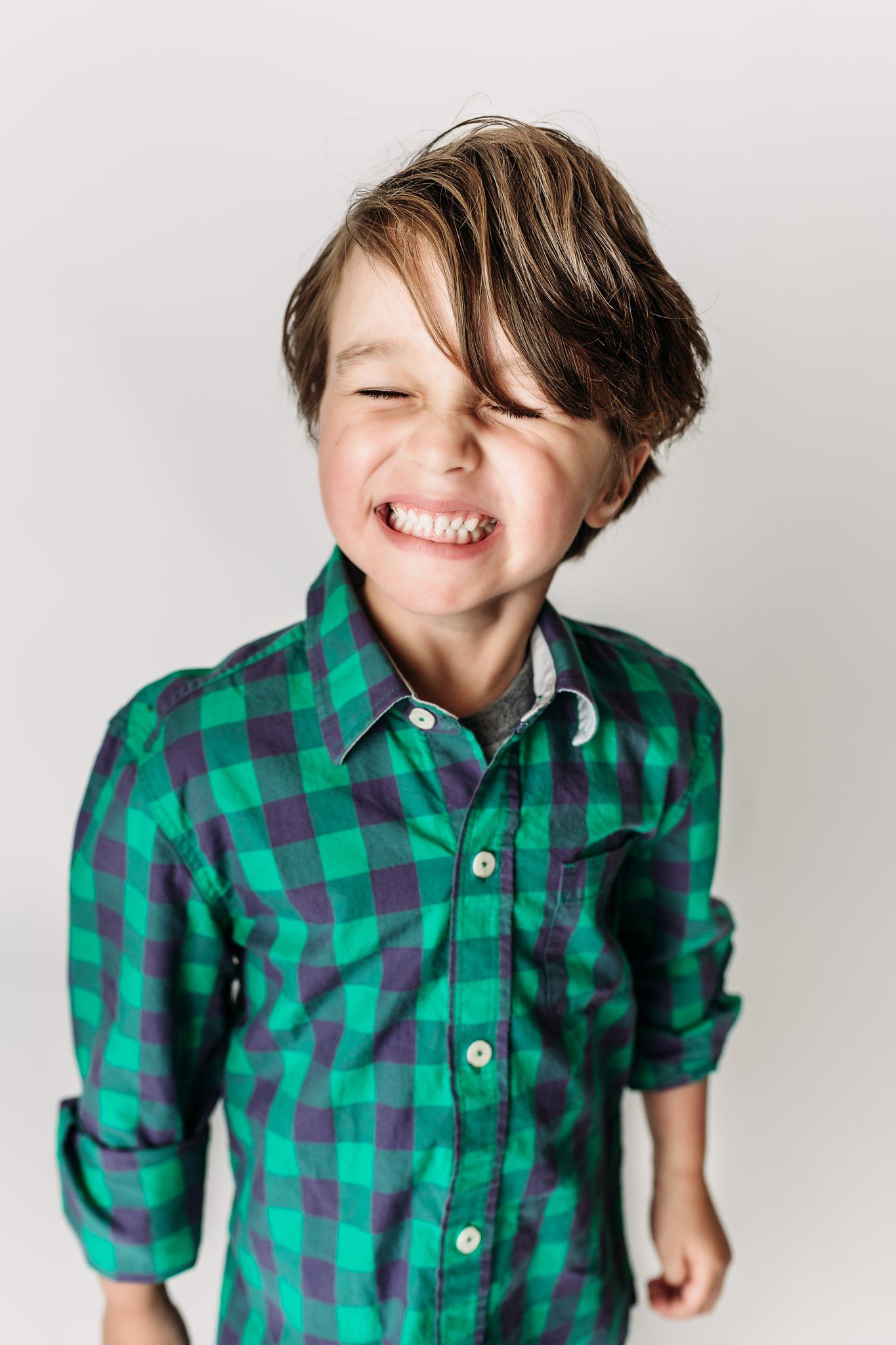 Child wearing green plaid shirt smiling with eyes closed against white background.