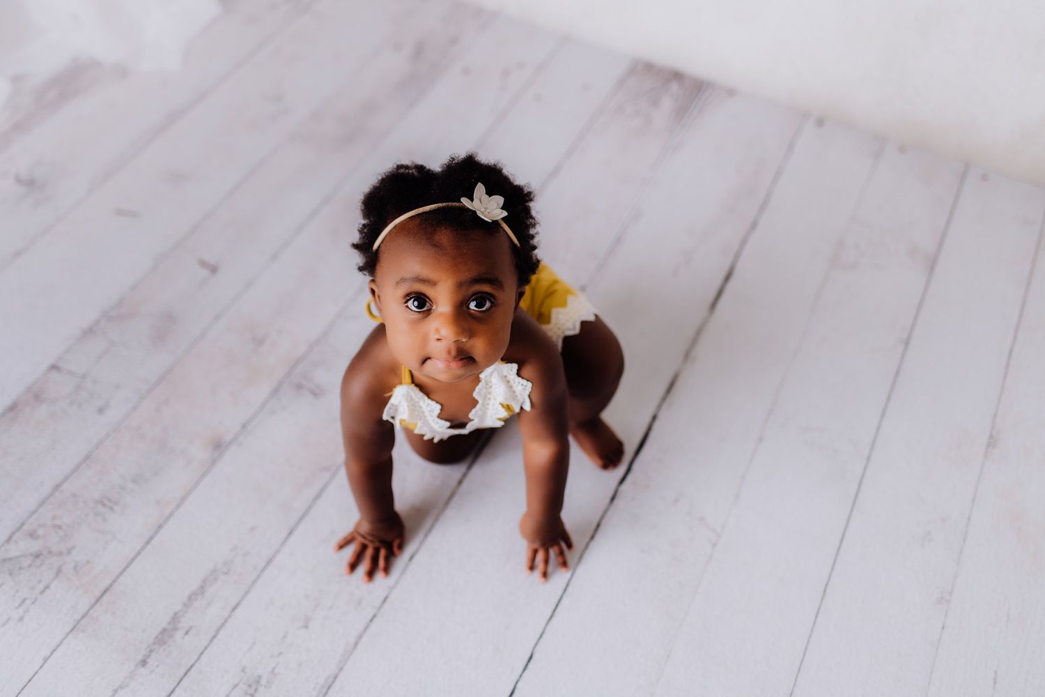 Baby in yellow outfit crawling on white wooden floor during photo shoot.