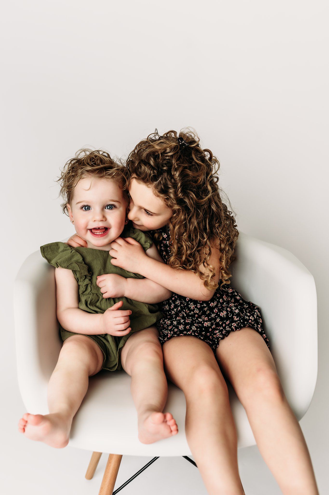 A heartwarming sibling moment captured on a white modern chair against a neutral backdrop.