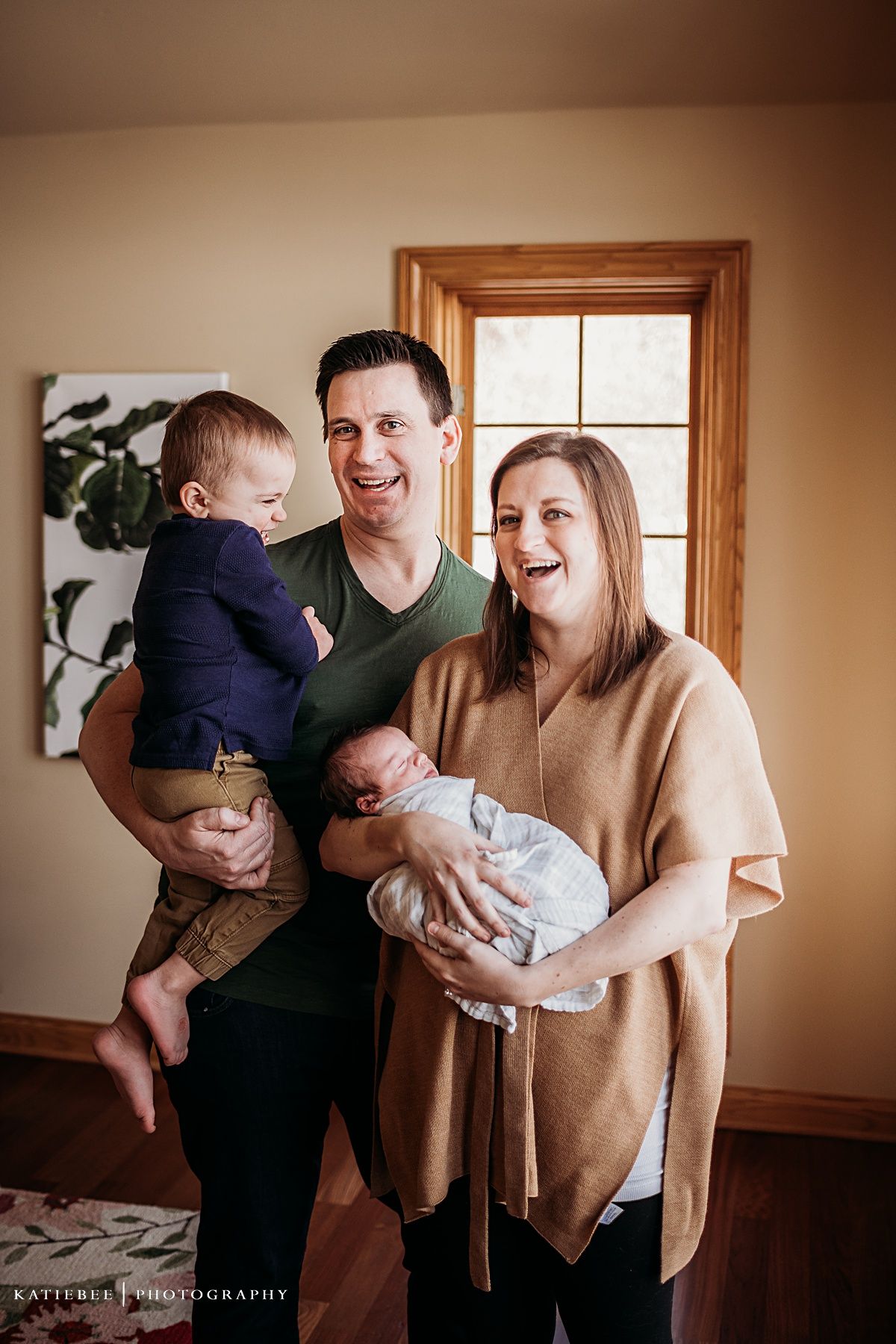 Portrait of a growing family with two small children in a sunlit room with wooden window frames.