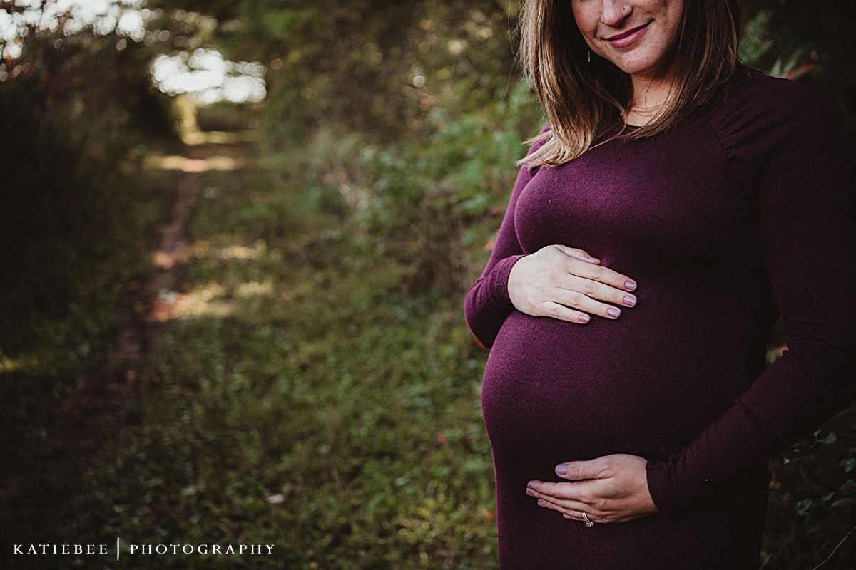 An expectant mother in a purple dress cradles her baby bump outdoors.