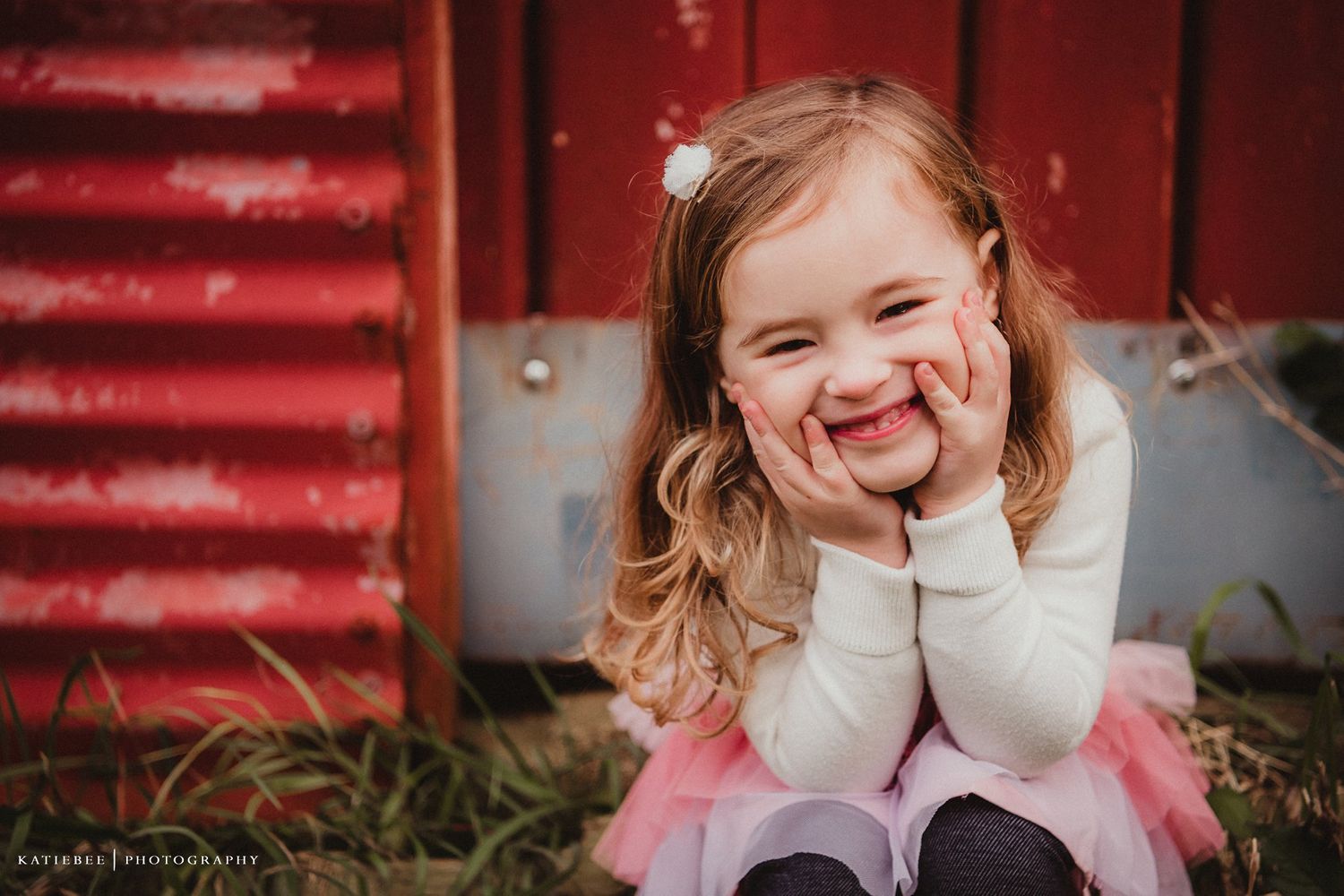 A joyful child in a white sweater laughs with hands on cheeks against red barn.